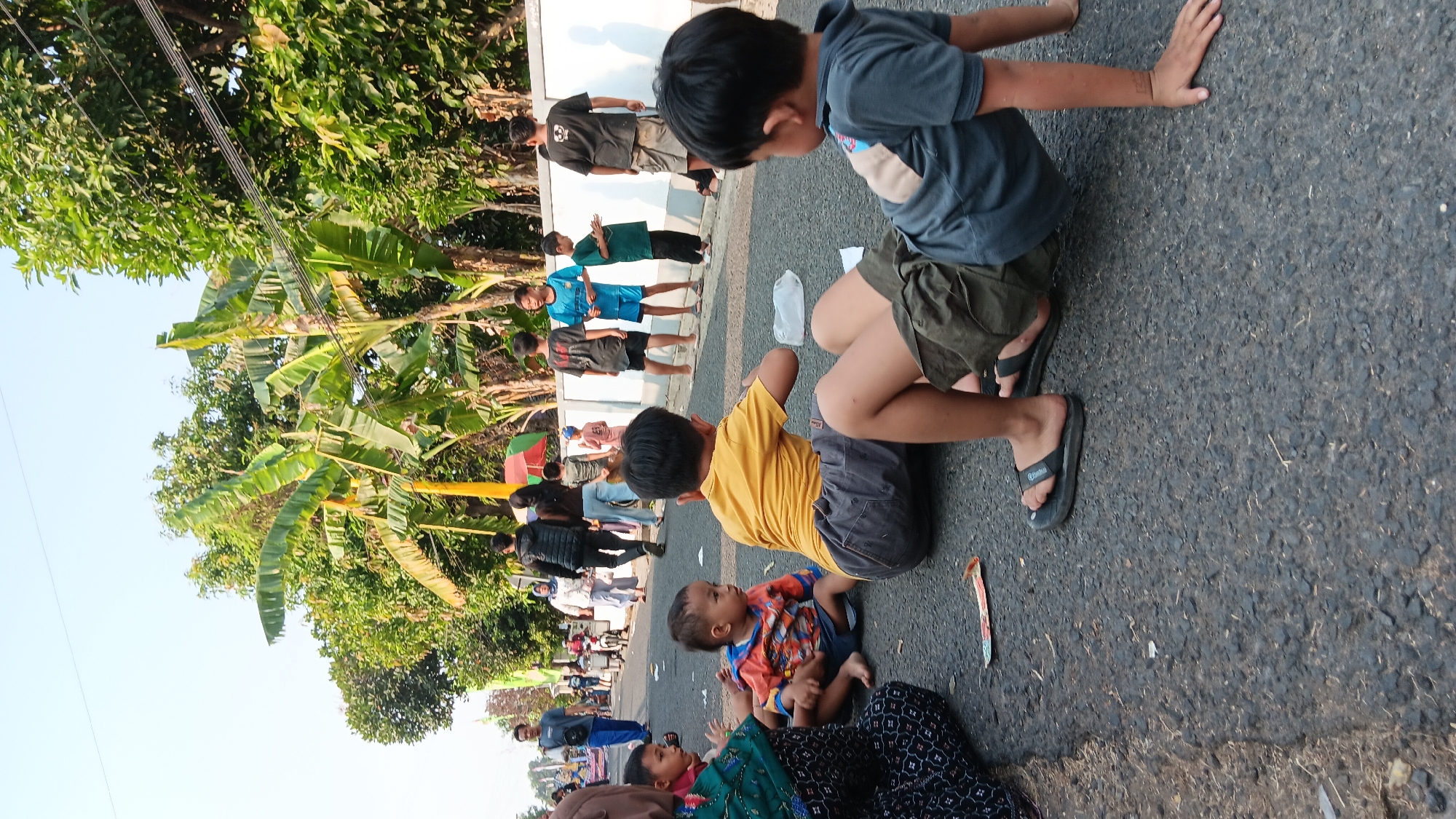 The image depicts an outdoor scene on a paved road, likely in the city of Cerme, Indonesia, given the context. Several individuals, predominantly children, are present. In the foreground, two young boys are sitting on the ground. One boy, wearing a yellow t-shirt and gray shorts, is facing away from the camera. To his right, another boy in a gray shirt and olive green shorts is also seated, leaning forward. Further to the left, a toddler in an orange and blue patterned shirt and dark shorts is sitting on the road. A woman, partially visible, wearing a patterned headscarf and a dark patterned dress, is seated next to the toddler.
In the mid-ground, a group of children and adults are visible. Some children are standing or walking, dressed in casual attire. There are trees and foliage lining the side of the road, with banana trees being prominent. A white wall runs along the side of the road, behind which more trees are visible. In the background, a man in a blue shirt and pants walks away from the camera, and other figures and a motorcycle are also present further down the road. The sky is clear and bright, indicating daylight. The ground is asphalt, with some debris and a piece of colorful paper visible near the foreground subjects. The overall impression is of a casual gathering or event taking place on a street.