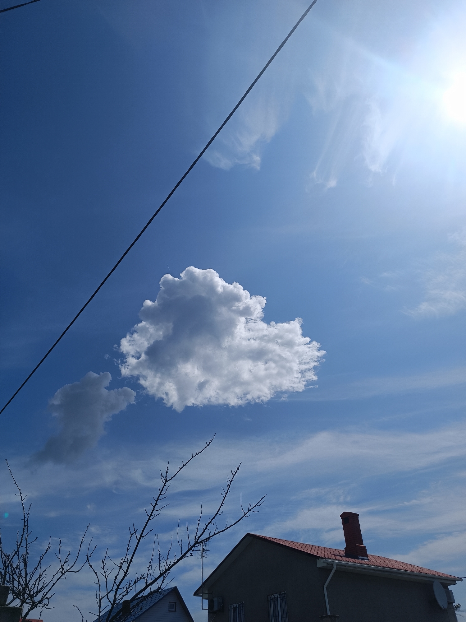 An overhead, low-angle shot shows a bright blue sky with scattered white clouds. A single black power line runs diagonally from the top left to the bottom center of the frame. In the upper right, the sun is intensely bright and partially obscured by a thin layer of white clouds. A large, puffy white cloud is centered in the middle of the sky, with a smaller, darker cloud situated to its left. In the foreground, the bare branches of a tree extend from the bottom left corner upwards. The roof and upper portion of a building are visible in the lower right, featuring a red roof, a brick chimney, and a satellite dish on the side. The sky is clear and cloudless except for the clouds depicted.