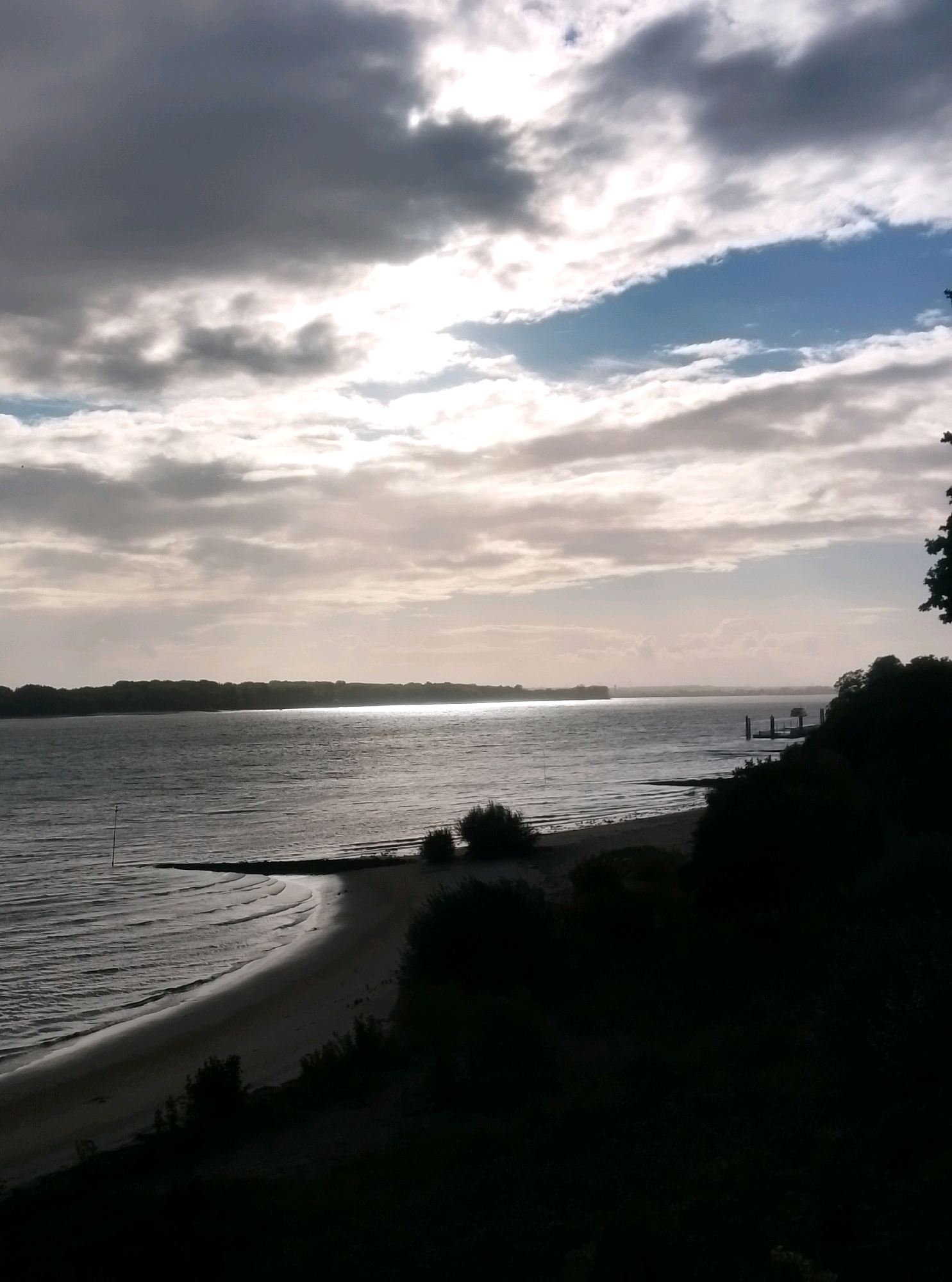 This image depicts a wide expanse of water, likely the Elbe River, in Wedel, Germany. The scene is characterized by a dramatic sky filled with both dark, heavy clouds and bright, illuminated patches, suggesting an approaching or receding storm. A calm body of water stretches across the middle ground, reflecting the sky's light. In the foreground, a sandy beach slopes down towards the water, bordered by dark, silhouetted trees and vegetation. A single, thin pole stands vertically in the shallow water near the beach. In the distance, a tree-lined horizon is visible, with a small pier and a few structures on the right bank of the river. The overall impression is one of a serene, natural landscape under a dynamic sky.