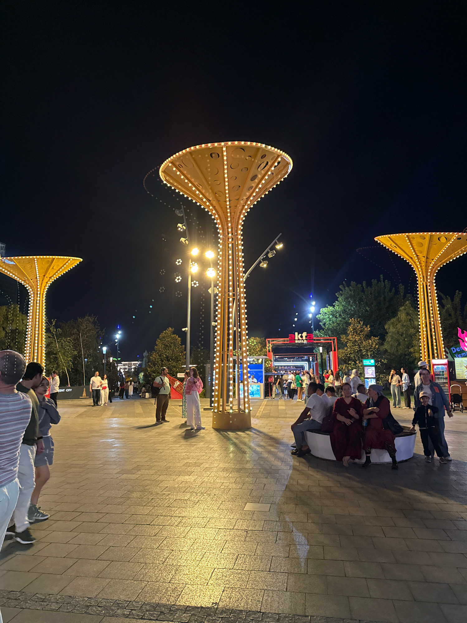 The image depicts a nighttime scene in a pedestrian area in Tashkent, Uzbekistan, likely a public square or park. Several prominent, illuminated structures, resembling stylized trees with broad, light-adorned canopies, dominate the foreground and midground. These structures are outlined with numerous small, warm-toned lights, creating a festive and illuminated atmosphere.
In the midground, there are various individuals engaged in different activities. Some are walking, while others are seated on a curved bench. A group of people are gathered near a building with signage that reads "akfa BUILD". There are also food or drink stalls visible in the background, suggesting a commercial or entertainment area. Trees line the walkway, their foliage silhouetted against the dark night sky. The ground is paved with large, textured tiles that reflect the ambient light. The overall impression is that of a lively public space during an evening event or gathering.