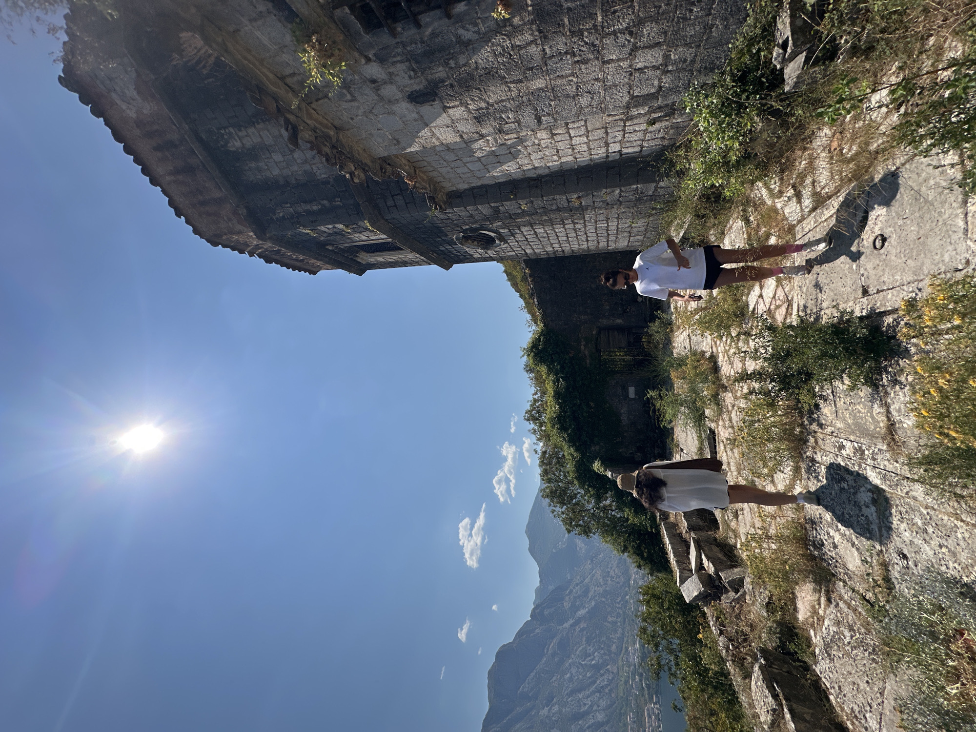 The image depicts two women walking on a stone path near a weathered, ancient stone structure, possibly a fortress or church, in Dobrota, Montenegro. The structure exhibits signs of decay, with crumbling stone and vegetation growing on its walls. The path is uneven, with scattered rocks and dry grass. In the background, a stunning panoramic view of the Bay of Kotor unfolds, with lush green mountains rising steeply from the turquoise water. The sky is a clear, vibrant blue, with the sun shining brightly, casting strong shadows.
The woman in the foreground, on the right, is wearing a white t-shirt, black shorts, sunglasses, and white sneakers with pink socks. She is walking towards the viewer's right and appears to be mid-stride. The woman in the background, on the left, is wearing a white long-sleeved shirt, shorts, and a straw hat. She is walking away from the viewer, towards the left, with a brown bag slung over her shoulder.
The overall scene suggests an exploration of a historical site with breathtaking natural beauty. The weathered stone of the structure contrasts with the vibrant blue of the sky and water, creating a dramatic and picturesque setting.