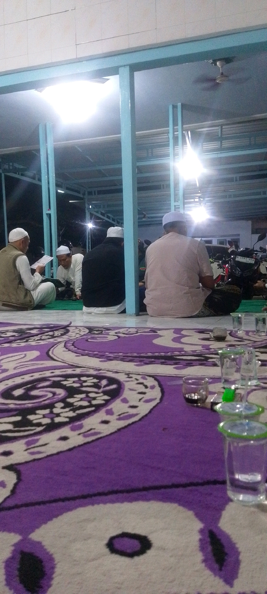 The image depicts a group of men seated on a purple and white patterned carpet, likely in an outdoor or semi-outdoor setting. The men are wearing traditional Islamic attire, including white skullcaps. One man on the left is reading from a book. In the background, there are light fixtures and architectural elements that suggest a covered area, possibly a mosque or a communal gathering space. The presence of motorcycles parked in the background indicates an urban or semi-urban environment. Several glasses are visible on the carpet, some containing dark liquid, suggesting a social gathering. The lighting is bright, casting shadows and highlighting the textures of the carpet and clothing.