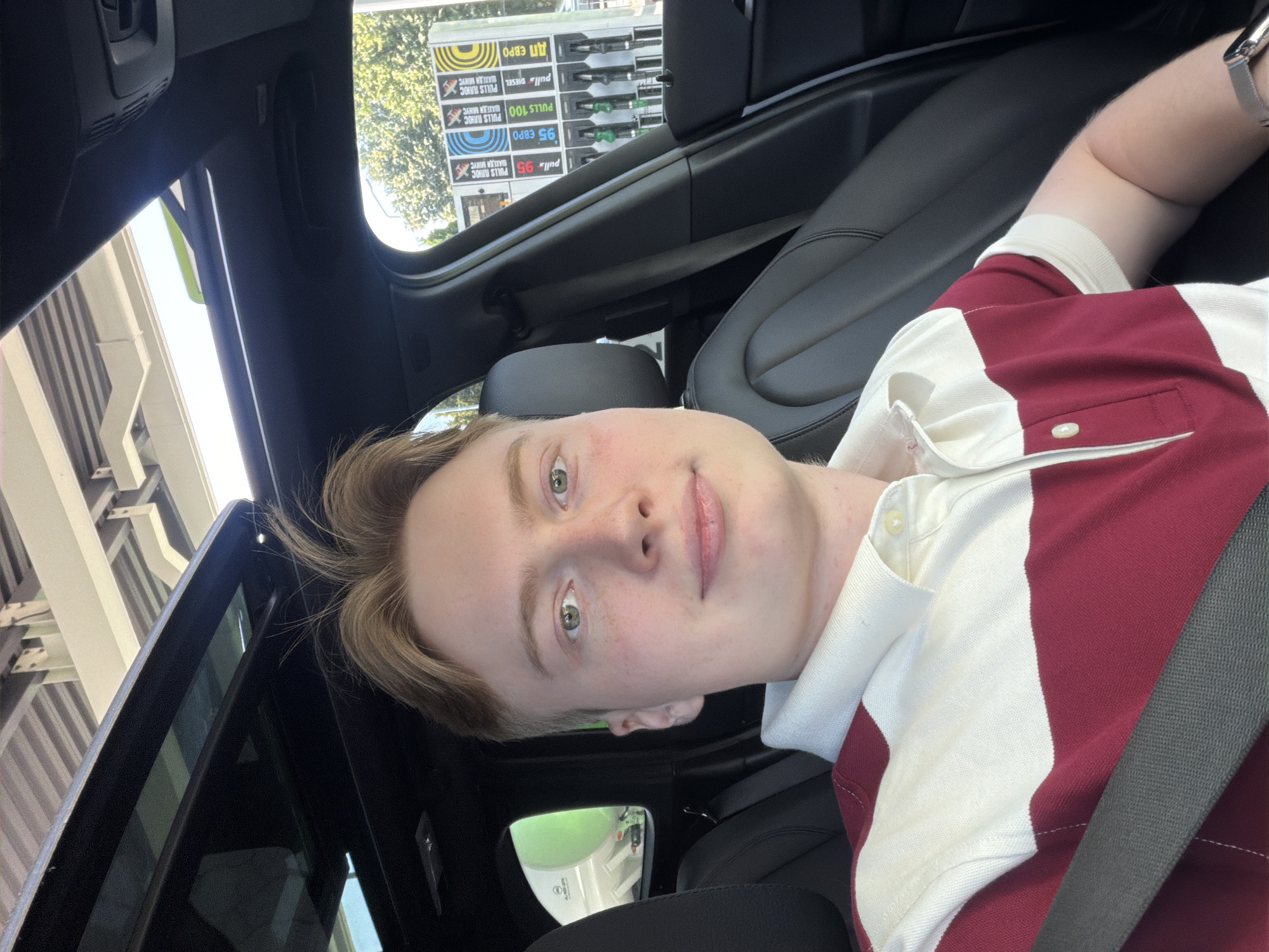 A young man with blond hair is sitting in the driver's seat of a car, looking directly at the camera. He is wearing a red and white striped polo shirt. The car's sunroof is open, revealing the sky and a partial view of a structure overhead, possibly a bridge or overpass. Outside the car, a gas station is visible with several fuel pumps. The gas pumps have various prices displayed, including "95 EURO" and "100". The scene suggests the man is at a gas station in Kyiv, Ukraine.