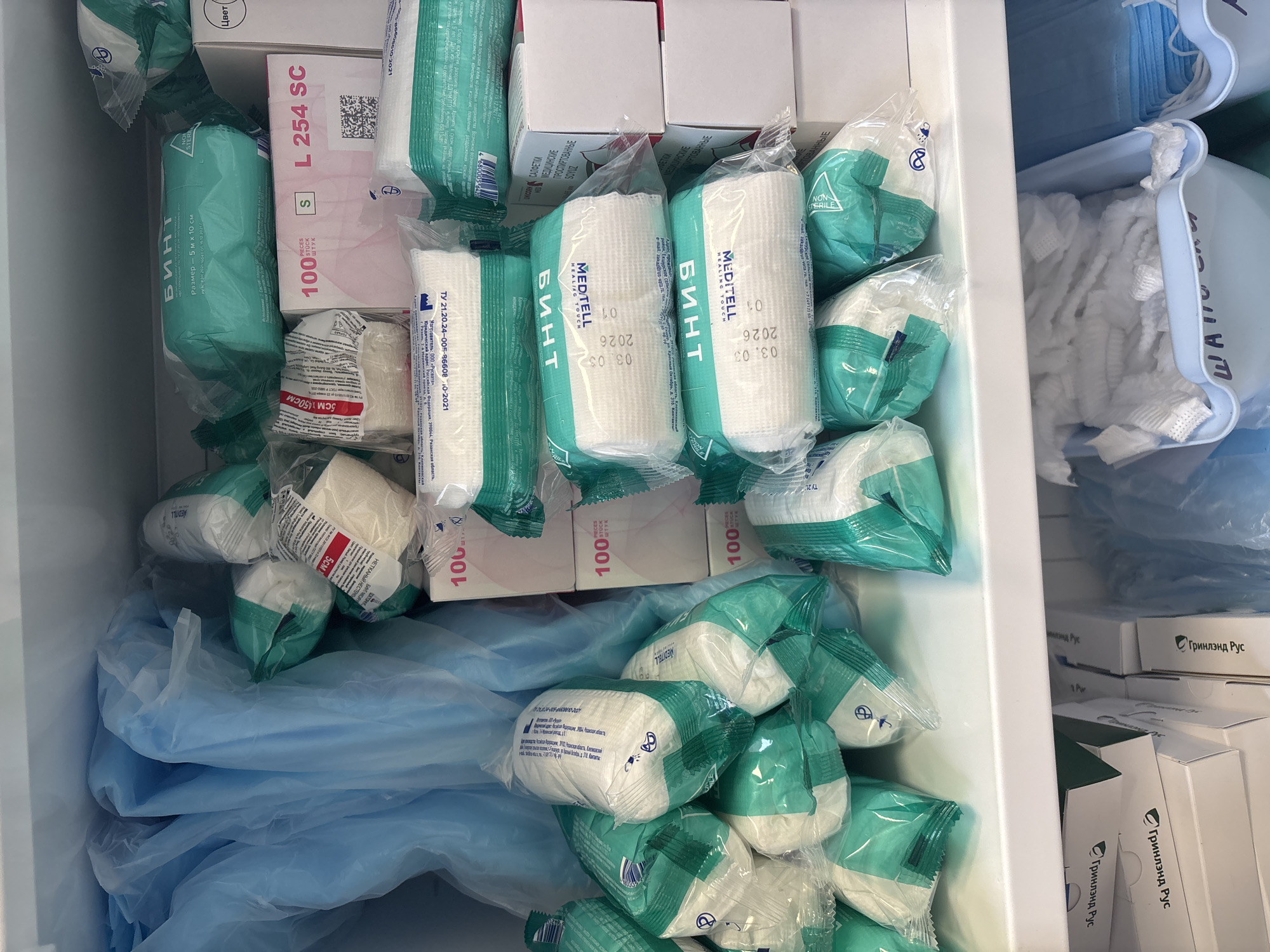 The image shows a shelf filled with medical supplies, primarily bandages and wound care products, likely in a storage room or pharmacy. The items are predominantly packaged in green and white wrappers, with some in blue plastic bags. Several boxes are visible with Cyrillic text, indicating they are of Russian origin. Some products are labeled with sizes, dates, and company names, such as "БИНТ" (bandage) and "MEDTELL HEALING TOUCH". In the lower portion of the image, a blue bin contains what appears to be crumpled gauze or bandages, and another bin holds blue masks. The environment appears to be a sterile or organized storage space.