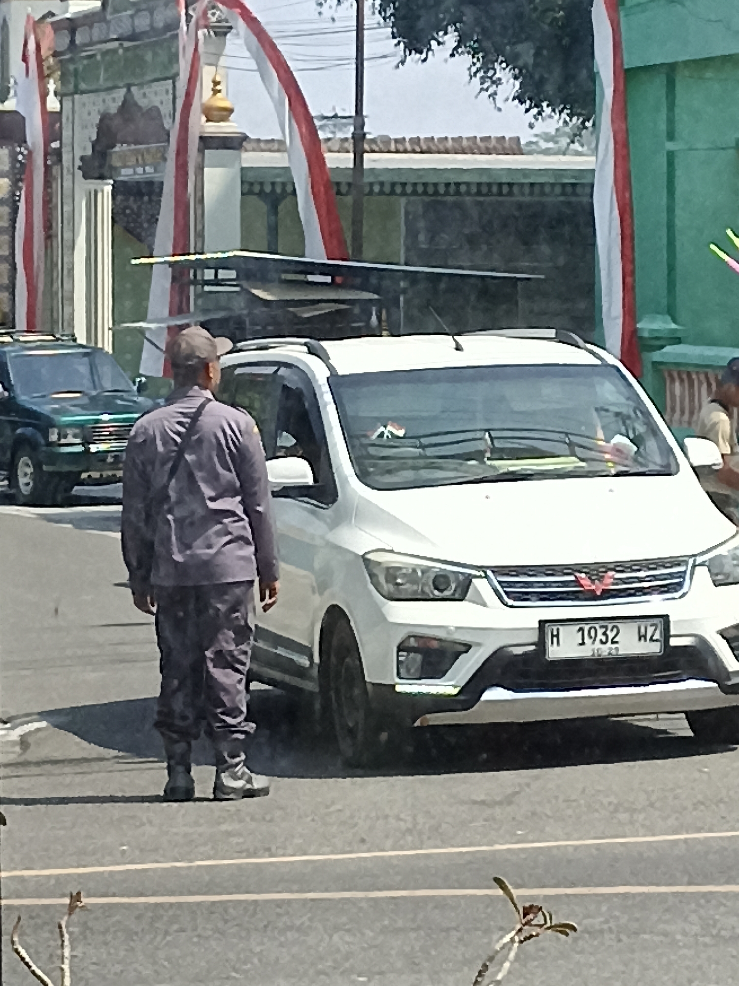 A man in a grey uniform and cap stands on the right side of a paved road, facing a white multi-purpose vehicle. The man is facing away from the camera, with his left shoulder slightly forward. He is wearing dark grey pants and a dark grey long-sleeved shirt, with a dark strap across his chest. He has black boots on. The white vehicle is facing the right of the image, with its front end clearly visible. A license plate on the front of the vehicle reads "H 1932 WZ". Red and white Indonesian flags are draped on poles in the background, attached to a building with green accents. A dark green SUV is visible on the left side of the road, further down the street. The ground is asphalt, with a white line marking the center of the road. The sky is bright and overcast. The scene appears to be in the city of Pulisen, Indonesia, given the presence of the Indonesian flags.