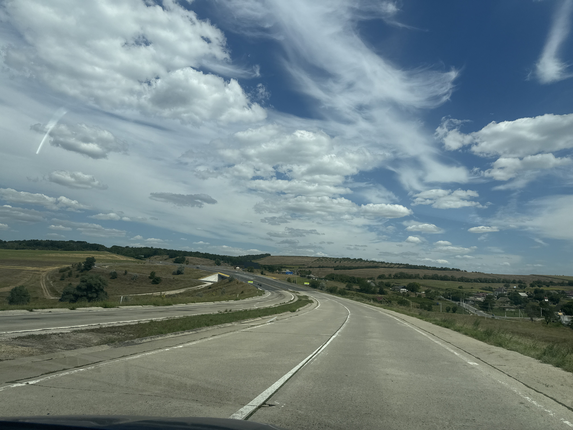 The image depicts a sunny day with scattered clouds over a rural landscape. A paved road, likely a highway or a major rural road, curves through rolling hills. The road has white lane markings. To the left of the road, there are grassy fields and some trees. Further in the distance, a small village is visible nestled among the hills, with scattered houses and trees. The sky is a vibrant blue with wispy white clouds. The scene appears to be a scenic view from a vehicle traveling on the road. The location is identified as Sagaidacul Nou, Moldova.