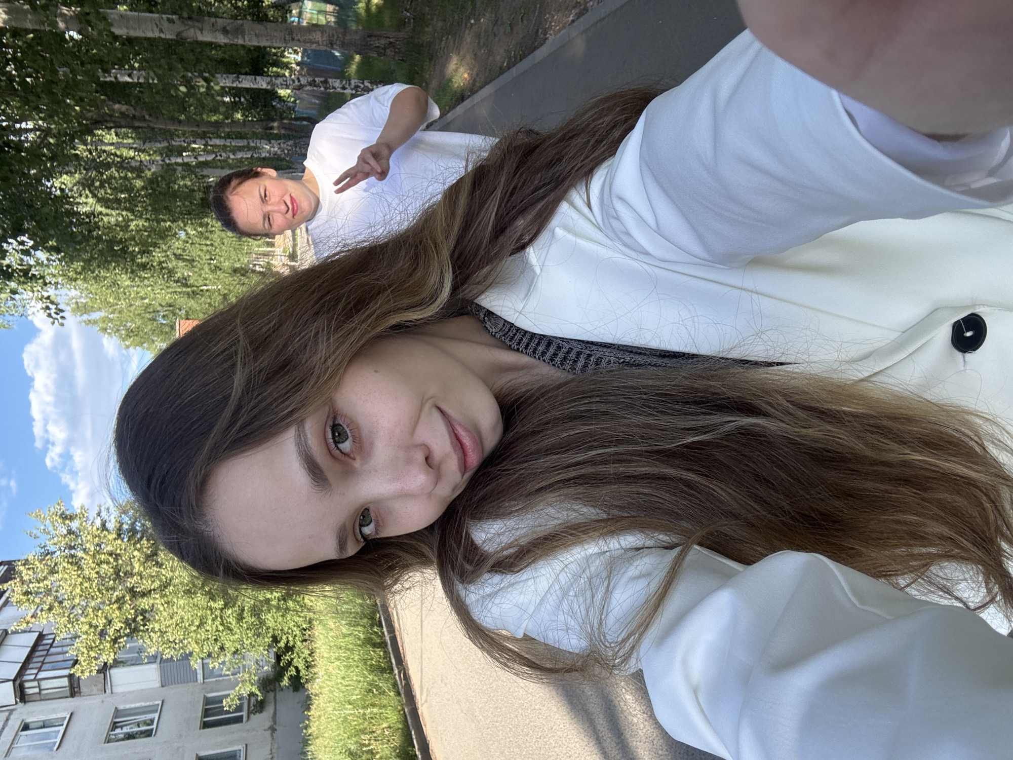 The media file is a selfie taken outdoors, with two people visible. The person in the foreground is a young woman with long, brown hair and green eyes, smiling at the camera. She is wearing a white jacket over a grey and black patterned top. The person in the background is a man wearing a white t-shirt and making a peace sign with his fingers. He is smiling and appears to be playfully posing.
The scene is set in a park-like area with trees and a paved walkway. Behind the people, there is a building with balconies and windows, suggesting an urban or suburban residential setting. The sky is blue with some clouds, and the lighting indicates it is daytime, likely a sunny day. The overall mood is cheerful and relaxed, suggesting a casual outing. There is no visible text in the image. The location context is Noviy, Russia.