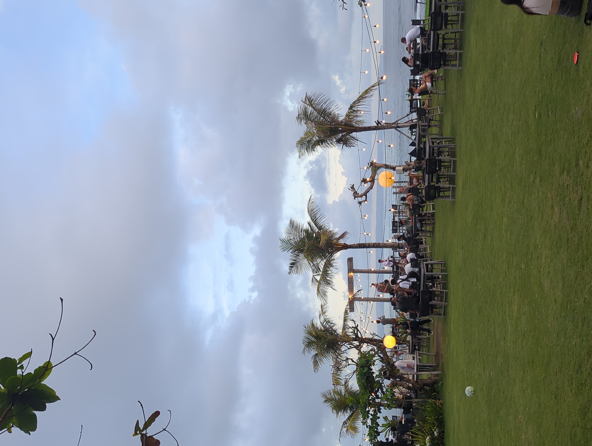 The image depicts an outdoor seating area in Mengwi, Indonesia. Tables and chairs are arranged on a grassy lawn, with several people seated at the tables. Palm trees are interspersed among the tables, and string lights are strung overhead. The sky is cloudy, and the ocean is visible in the background. A soccer ball sits on the lawn near the lower right edge of the image.