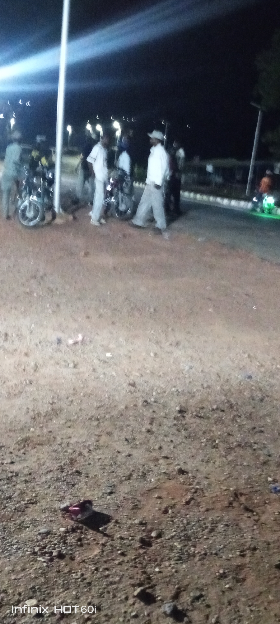 At night in Adankolo, Nigeria, a group of men congregate by motorcycles near a light pole. The ground is dirt and gravel. A shoe lies discarded on the ground. In the background, there's a road, possibly a building, and more lighting.