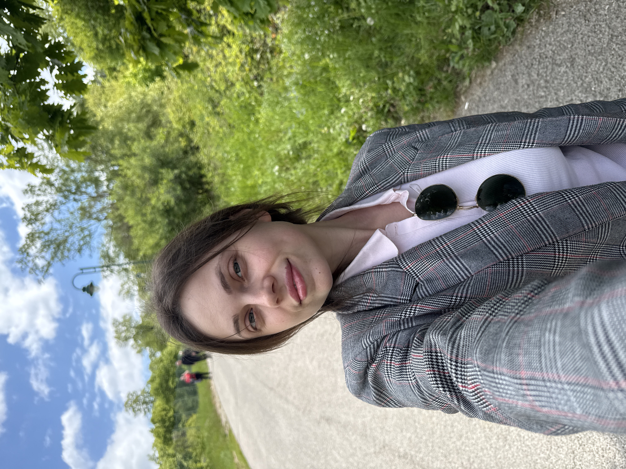 A woman is posing for a selfie in what appears to be a park in Sremska Kamenica, Serbia. She is wearing a gray plaid blazer over a light pink shirt and sunglasses are hooked on her shirt. Behind her is a paved path flanked by grass and trees. Further in the background, there are other people walking on the path under a blue sky with clouds. A lamp post can also be seen in the background.