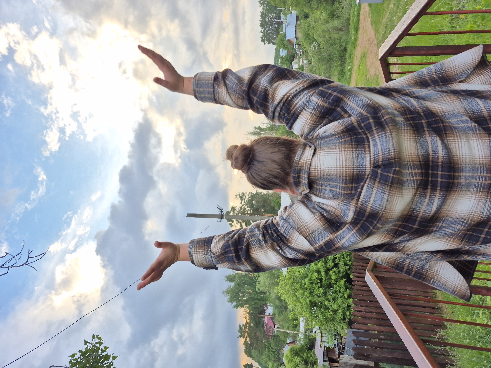 This outdoor photograph, captured in Chaikovsky, Russia, shows a person viewed from behind, their arms stretched upwards and outwards towards a bright, partly cloudy sky. The individual is wearing a brown, white, and black plaid shirt, and their hair is tied in a bun. The pose suggests a moment of joy, freedom, or appreciation for the expansive view.
The upper portion of the image is dominated by a dynamic sky, featuring both clear blue patches and soft, illuminated clouds where the sun appears to be breaking through, casting a warm glow. This light indicates either a late afternoon or early evening setting.
In the midground and background, a lush green landscape unfolds, characterized by numerous trees and dense foliage. A dirt path is visible, winding through the greenery, and several distant houses or structures are nestled amidst the trees, suggesting a rural or semi-rural environment. A wooden fence and a reddish-brown metal railing are prominent on the right side of the frame, indicating the person is standing on an elevated area such as a porch or balcony. A power pole with wires is also visible against the sky in the distance. There is no visible text in the image.