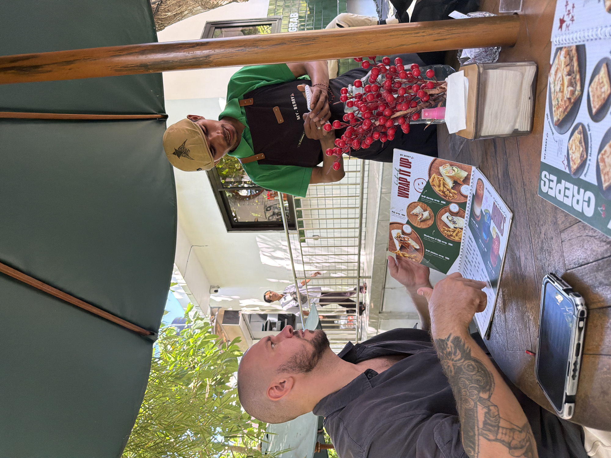 In Kuta Selatan, Indonesia, a customer with a shaved head and beard, wearing a dark grey shirt and tattoos on his left arm, sits at a wooden table under a large dark green umbrella. He is looking at a menu and gesturing with his right hand. A waiter, wearing a green shirt and a black apron with the logo "MY.BERRY", stands to the right, holding a pen and notepad, appearing to take an order. A red berry arrangement in a vase is placed on the table, along with a tissue dispenser. In the background, a white building with windows and a railing is visible, with another person walking on a walkway. The scene suggests an outdoor dining setting at a cafe or restaurant.