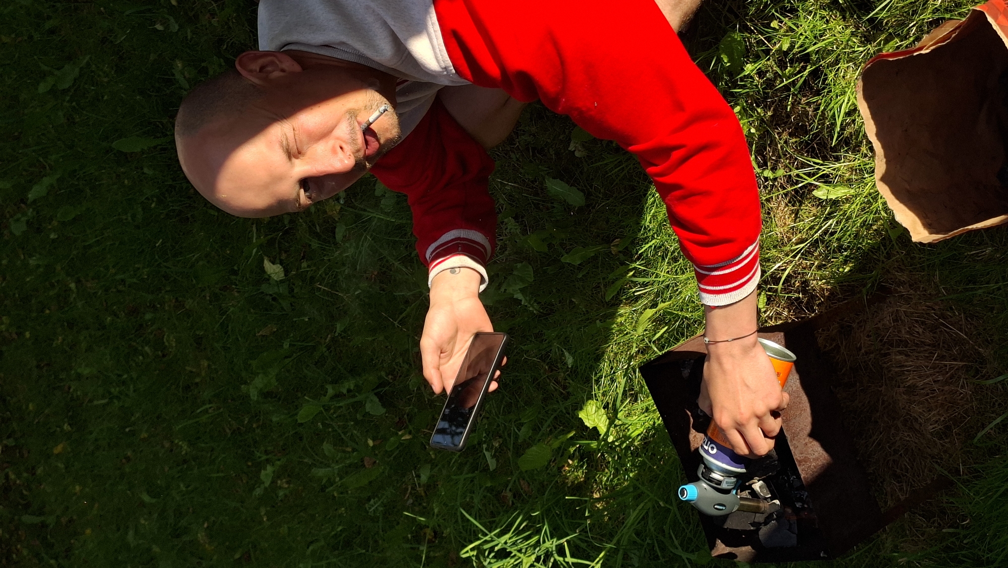 A bald man with a cigarette in his mouth is squatting on green grass, holding a smartphone in his left hand and a can with a lighter attachment in his right. The man is wearing a red sweater with white stripes on the sleeves and collar. He is looking at the camera and winking. In front of him is a metal grill with charcoal. A brown paper bag is visible in the bottom right corner of the image. The lighting suggests it is daytime, likely outdoors. The scene appears to be in a rural area, possibly a backyard or park, consistent with the location of Забалоцкі сельскі Савет, Belarus.