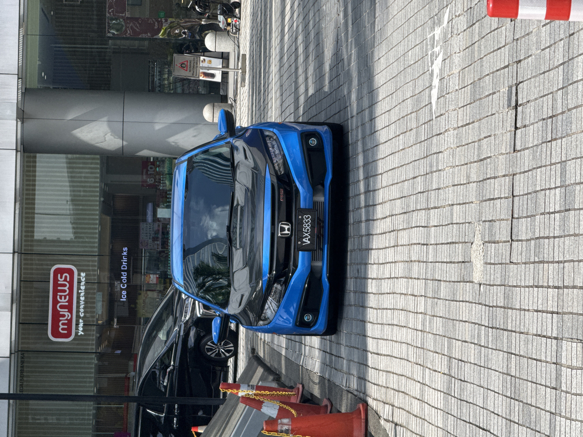 The image shows a blue Honda Civic RS parked in front of a myNEWS convenience store. The car's license plate reads "VAX5833". Several orange traffic cones are arranged near the vehicle. A black car is partially visible behind the blue car. The building's window displays "Ice Cold Drinks" and some prices. There is also a sign stating “NO PARKING. Wheel Clamping Zone. Release Fee RM100.00.” The surrounding area appears to be a paved walkway, likely in an urban setting. The reflections in the building's glass suggest other structures and vegetation nearby. Based on the "myNEWS" store, the location is likely Kuala Lumpur, Malaysia.