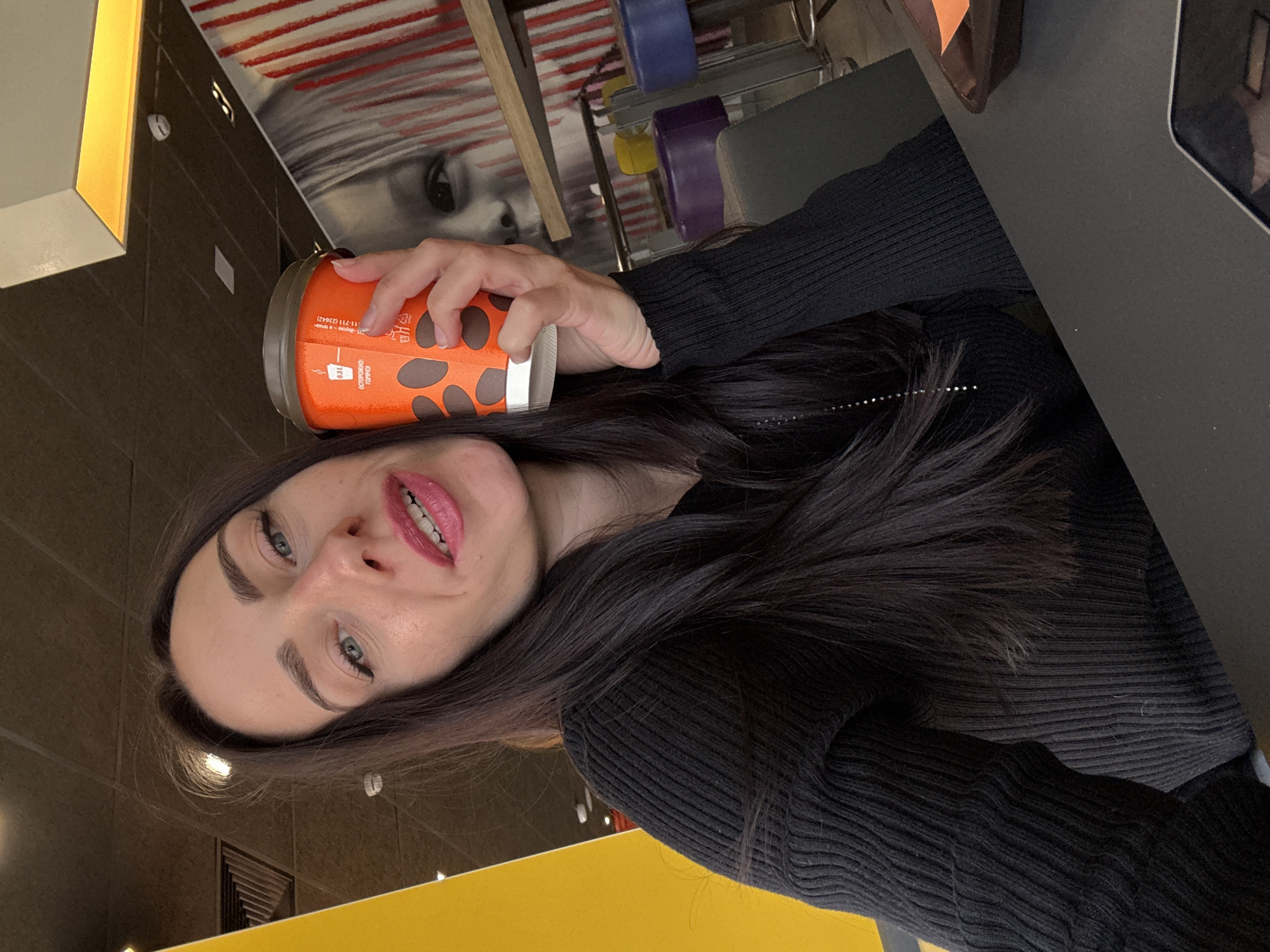 A woman with long dark hair, wearing a black sweater, holds an orange paper coffee cup. She has light blue eyes and red lipstick. The background shows a restaurant interior with a black and gray table, a painting of a woman's face, and colored chairs. The scene is likely set in a public space, possibly a café or restaurant. It is unknown if this is in St. Petersburg, Russia.