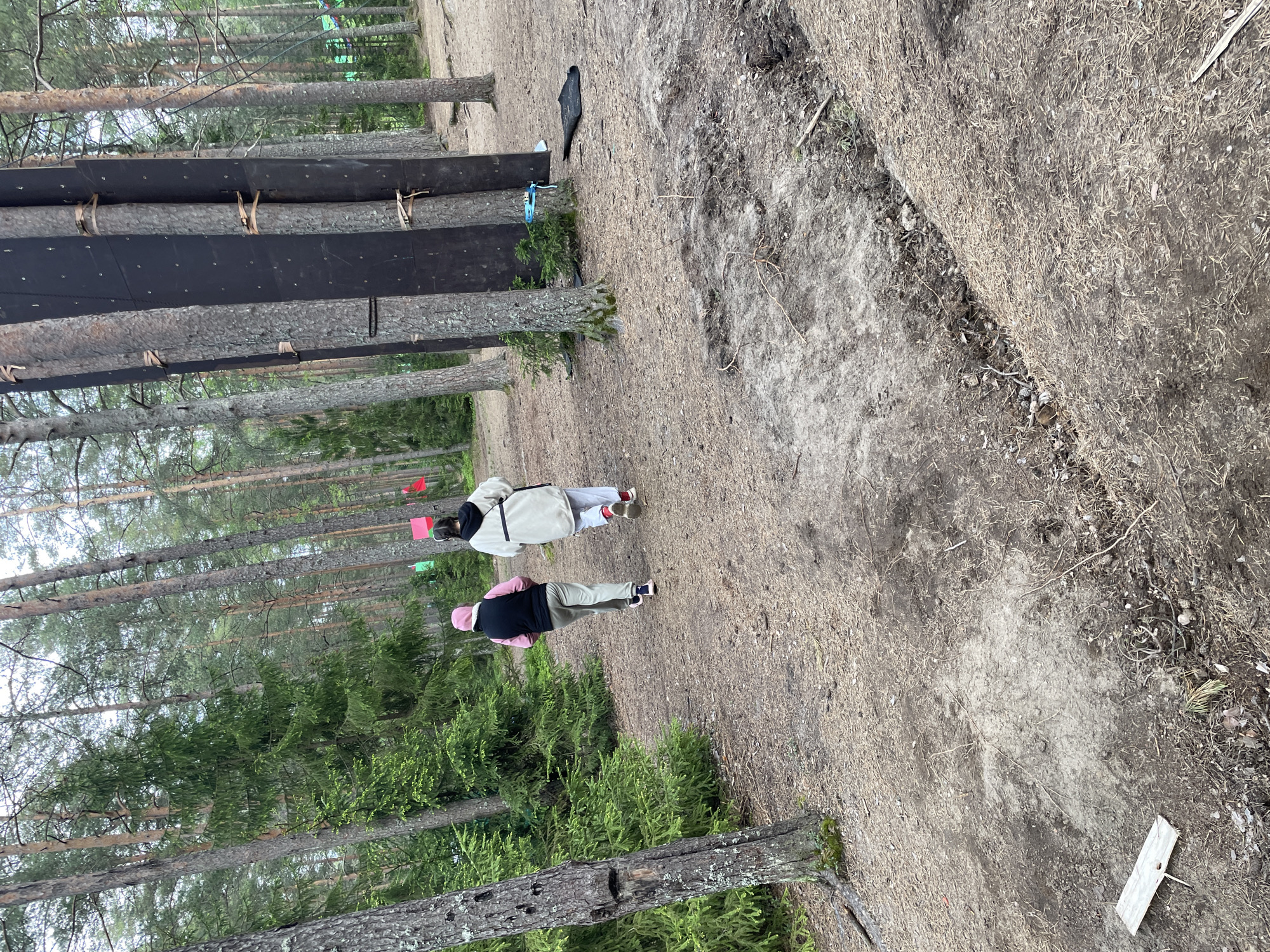 The image depicts a woodland scene near Unknown, Russia. Two individuals are visible, both wearing casual clothing. One wears a pink hat and the other appears to be wearing a dark jacket. They are walking on a path that appears to be covered with pine needles and dirt. The path is adjacent to a forest consisting primarily of pine trees, some of which are covered with dark rectangular panels. Other objects and debris are visible scattered on the forest floor.
