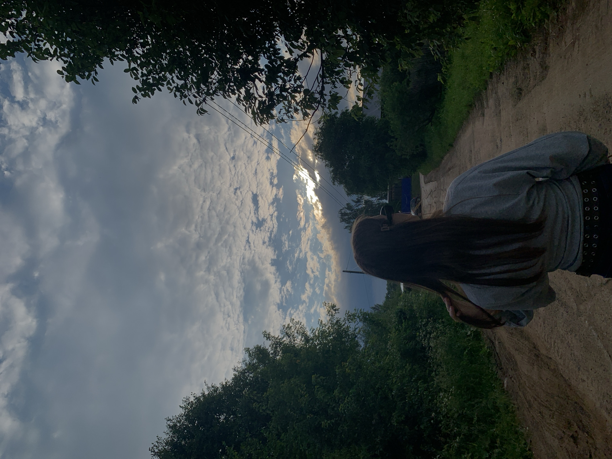 The image depicts a young woman from behind, standing on a dirt road in a rural area that appears to be located in Fokinsky District, Bryansk Oblast, Russia. The road is flanked by dense green foliage and trees on both sides, with a cloudy sky overhead. The woman has long, dark hair and is wearing a grey long-sleeved shirt and dark pants. She is looking down the road towards a distant point where the sun is peeking through the clouds, creating a dramatic lighting effect. Power lines are visible stretched across the sky. The overall scene suggests a peaceful, natural environment with a hint of moody weather.