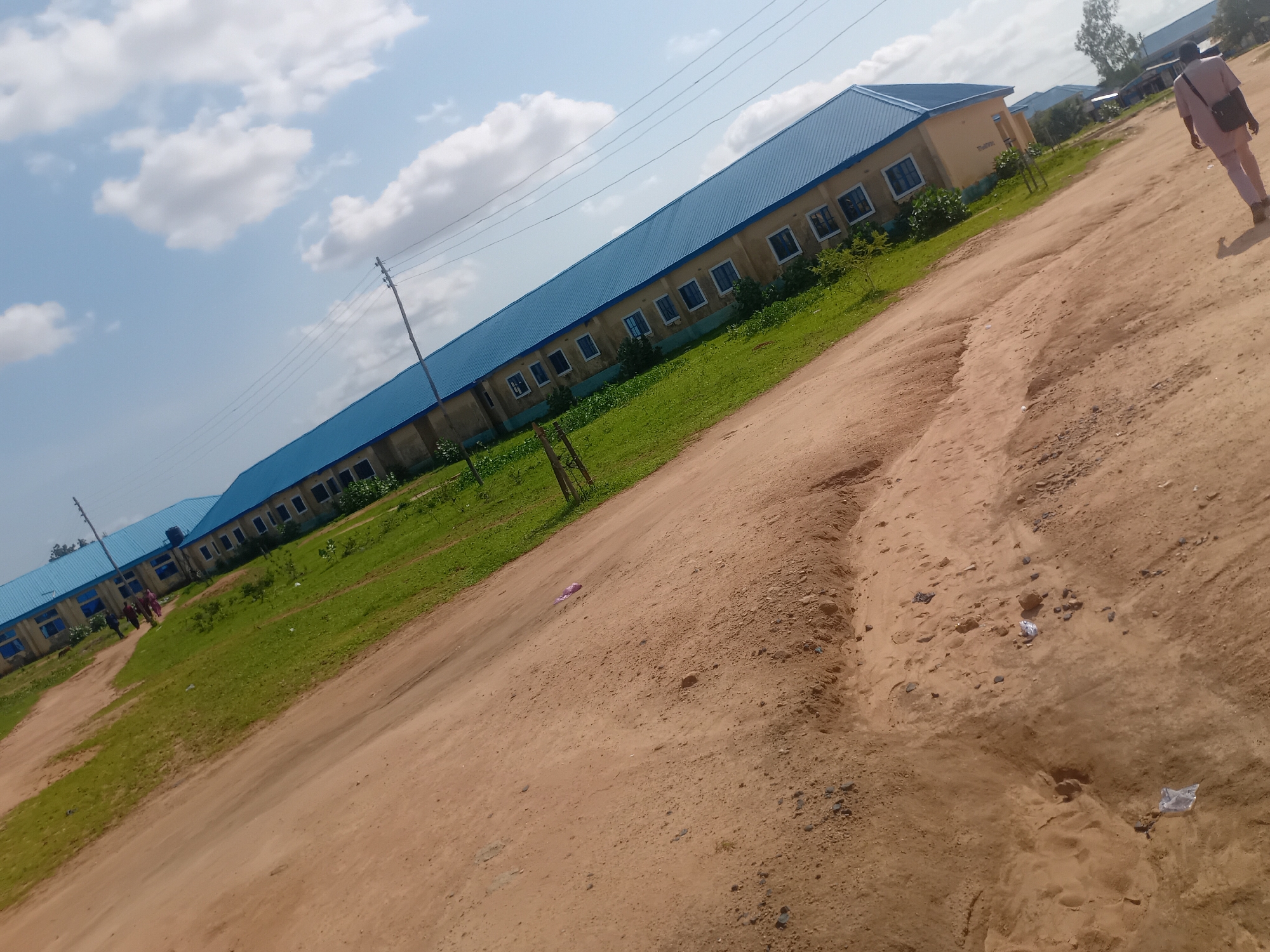 The image depicts a wide, unpaved dirt road running diagonally from the bottom right to the top left corner of the frame. The road surface is predominantly sandy with scattered rocks and some small depressions. To the left of the road, there is a strip of green grass, indicating a slightly lower elevation than the road. Beyond the grass, a long, light-colored building with a bright blue, corrugated metal roof stretches along the left side of the image. The building has numerous windows with dark frames. Several people are visible walking on the dirt path in front of the building, and one individual is walking away from the camera on the road in the foreground. In the background, more of the same building structure is visible, along with some trees and utility poles with power lines. The sky is partly cloudy with a bright, clear blue expanse. The overall scene appears to be an outdoor setting, possibly on the outskirts of a settlement or in a more rural area.