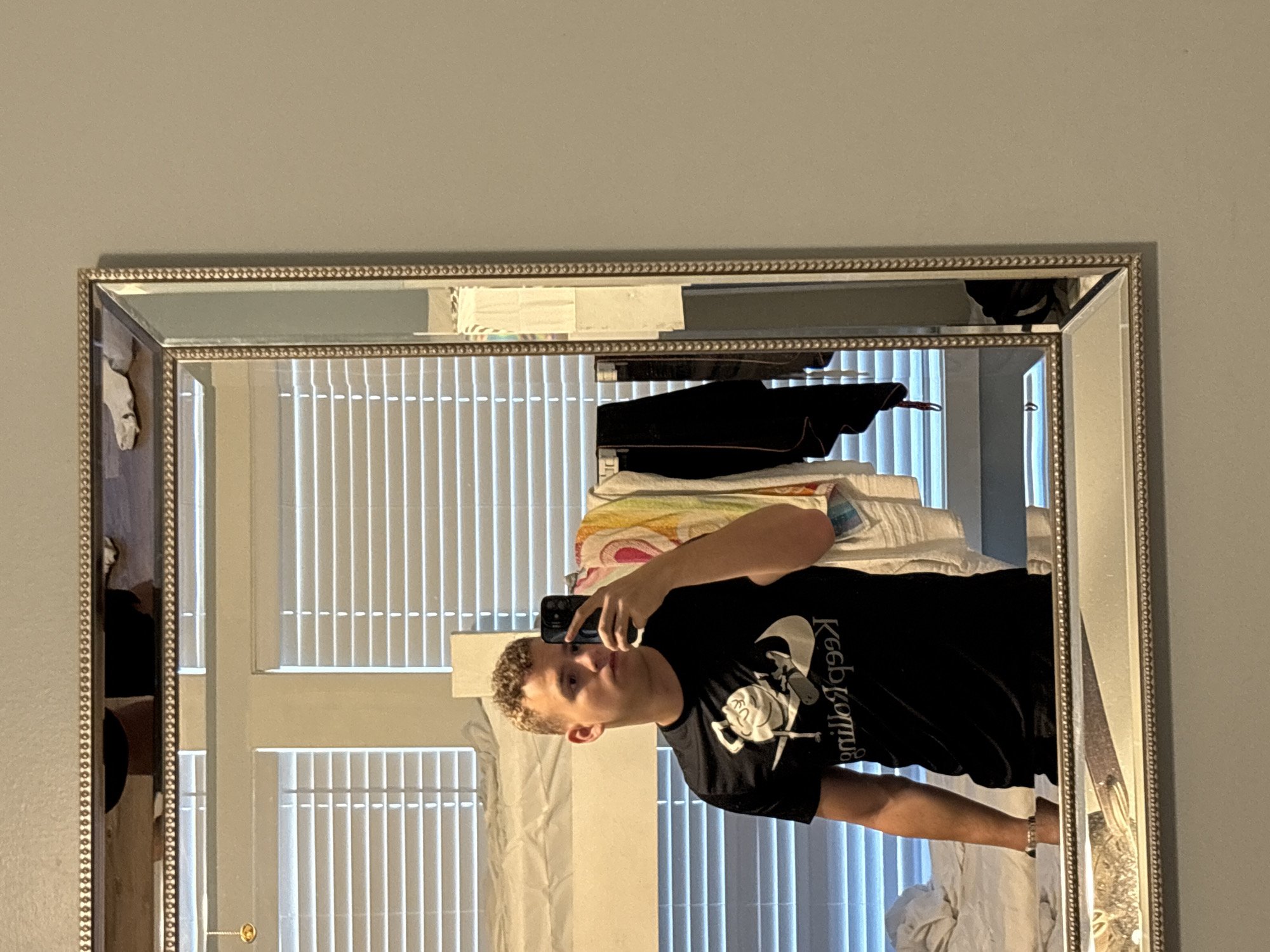 In Panama City Beach, Florida, USA, a young man with light brown, curly hair is taking a mirror selfie. He is wearing a black t-shirt with a graphic that appears to be a cartoon character and the text "Keep Rolling" in reverse. He is standing in what appears to be a bedroom, with blinds covering a window in the background. A bunk bed is visible to the left, and several articles of clothing are hanging on a rack to the right. The mirror has an ornate, beaded frame. The man is holding a black smartphone in his right hand to take the photo.