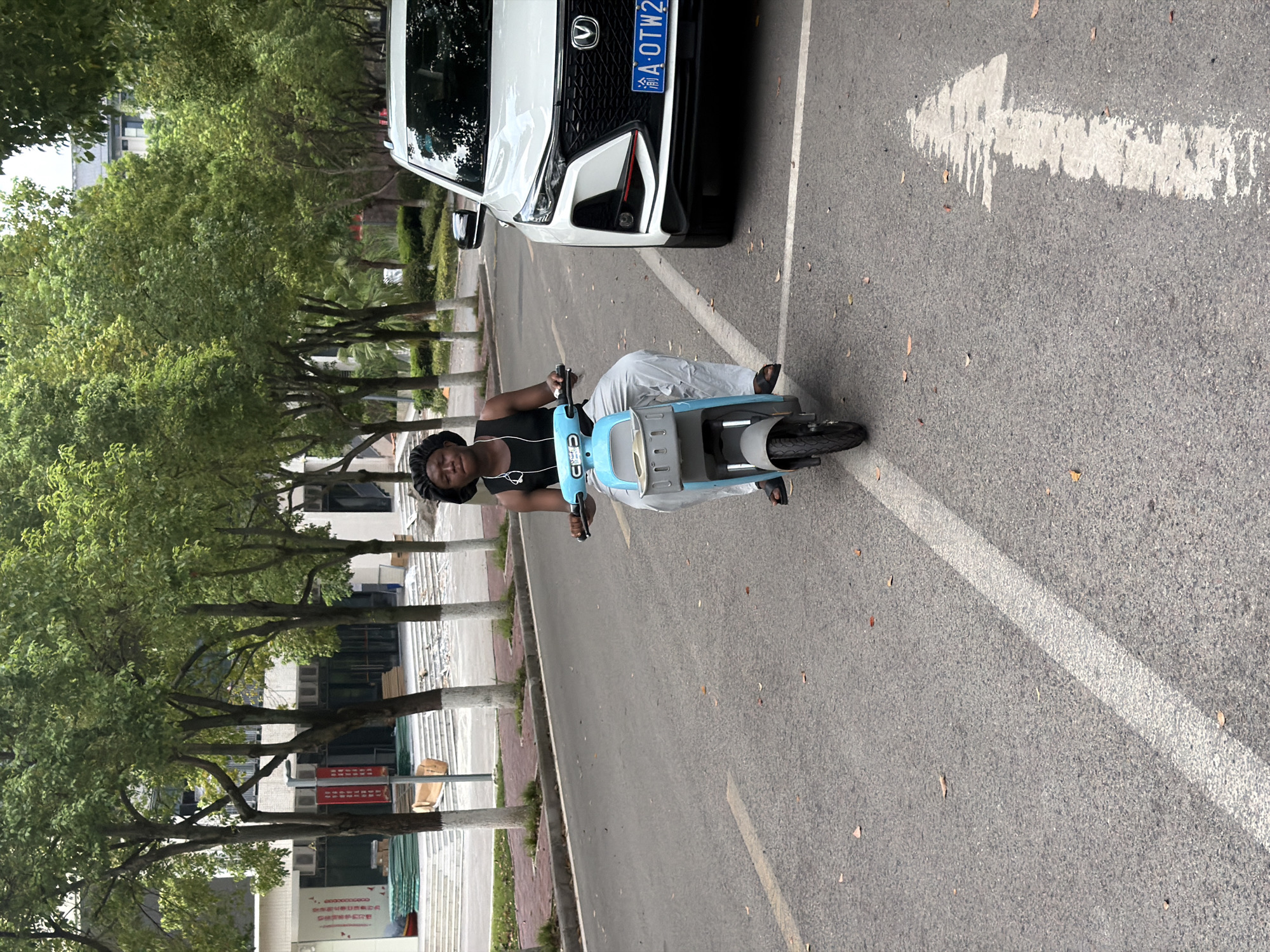 In Jiangjin, China, a young Black person wearing a black tank top, light-colored pants, and a dark head covering is sitting on a light blue electric scooter. They are wearing white earbuds and appear to be looking slightly off-camera. The scooter has a grey basket on the front. To the right of the scooter is the front of a white SUV with a blue license plate that reads "A OTW2". The scene takes place on an asphalt road with a white directional arrow painted on it, indicating traffic flow. In the background, there are rows of trees with green foliage and some buildings, one of which appears to be a large structure with white steps leading up to it. The overall environment suggests an outdoor urban or campus setting.