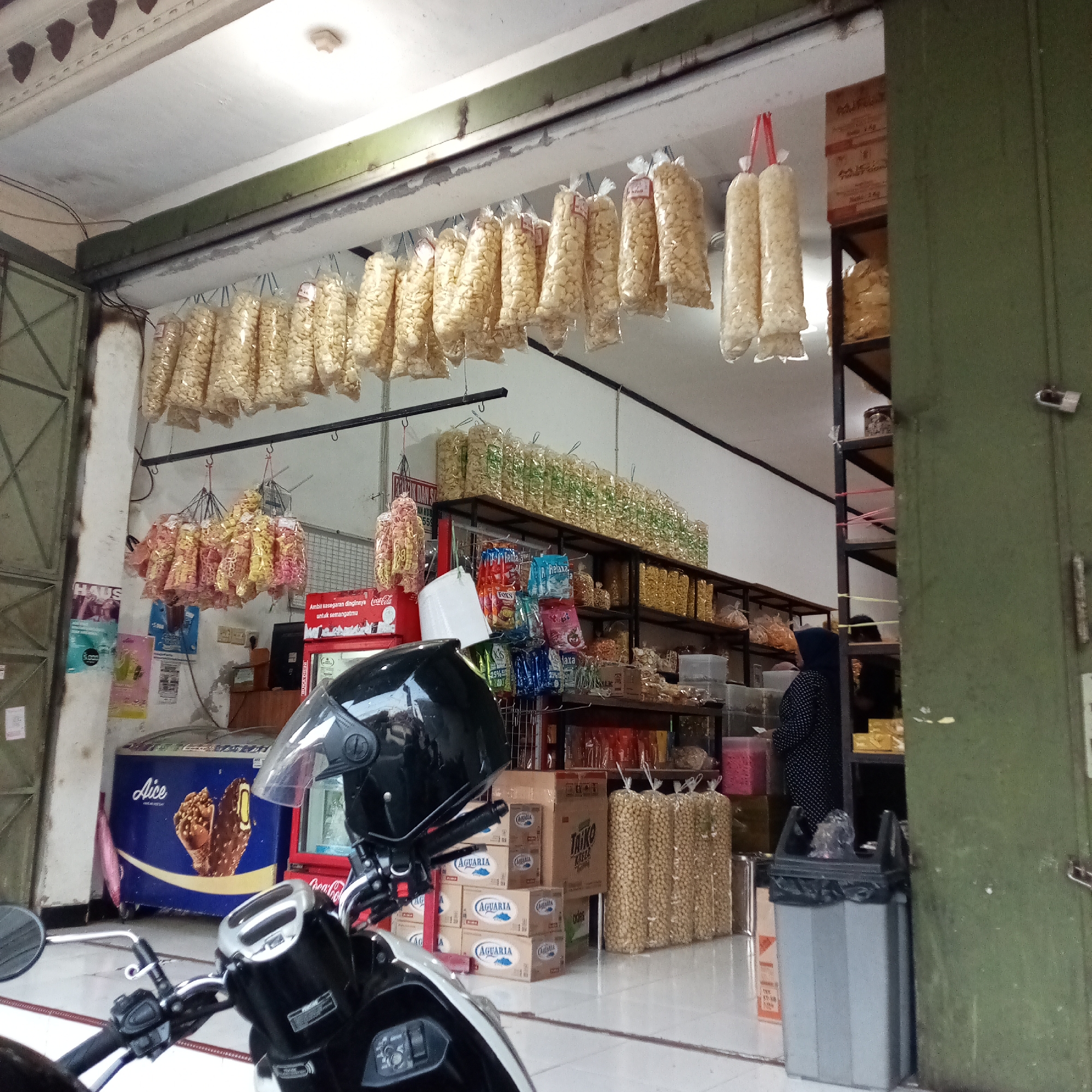 The image depicts the exterior of a small shop in the city of Sidoharjo, Indonesia. The shop is open to the street, with a green metal door on the right side of the frame. Large quantities of snacks are displayed, predominantly in clear plastic bags hung from the ceiling and on shelves. These snacks appear to be a type of puffed corn or cracker.
In the foreground, the handlebars and front portion of a black motorcycle with a black helmet are visible. The motorcycle is parked in front of the shop entrance.
Inside the shop, an ice cream freezer with an "Aice" logo and an image of an ice cream cone is prominent. Behind the freezer, a red Coca-Cola branded refrigerator can be seen. Shelving units filled with various packaged snacks and boxes are arranged along the back wall. A person wearing a dark headscarf and a patterned black and white outfit is partially visible inside the shop, interacting with items on a shelf. A trash can with a black liner is positioned near the entrance. The flooring appears to be tiled. Posters and signage are affixed to the wall. The overall impression is that of a local convenience store or snack vendor.