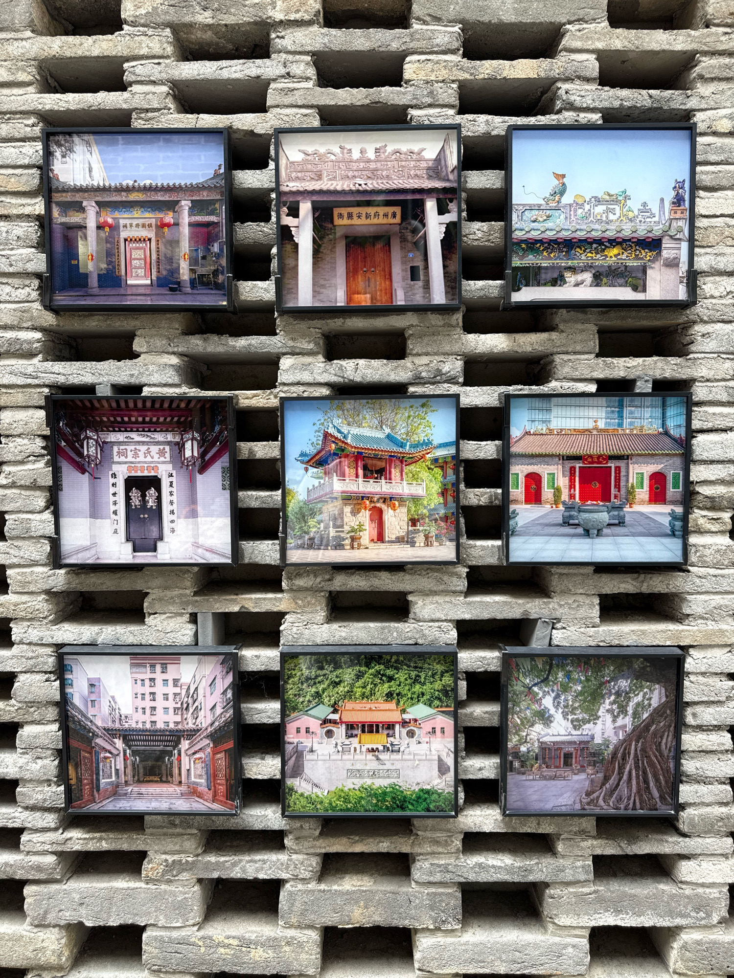 The image displays nine framed photographs mounted on a textured brick wall in Nanshan District, China. The photographs appear to be of traditional Chinese architecture, likely temples or ancestral halls, and are arranged in a 3x3 grid. The top row features three images: the first shows a building entrance with pillars and red lanterns, the second depicts a ornate rooftop with ceramic figures, and the third showcases a colorful temple rooftop with detailed decorations. The middle row contains three more architectural scenes: the first is an entrance to a courtyard with a prominent dark door and calligraphy, the second is a multi-story structure with red doors and a tiled roof, and the third is a courtyard with a large building, red doors, and two large incense burners in the foreground. The bottom row includes three images: the first is an alleyway with traditional buildings and apartments in the background, the second shows a temple complex on a hillside with a wide staircase, and the third features a courtyard with a large, gnarled tree in the foreground and a building in the background. The brick wall itself has a distinctive pattern of recessed rectangular openings.