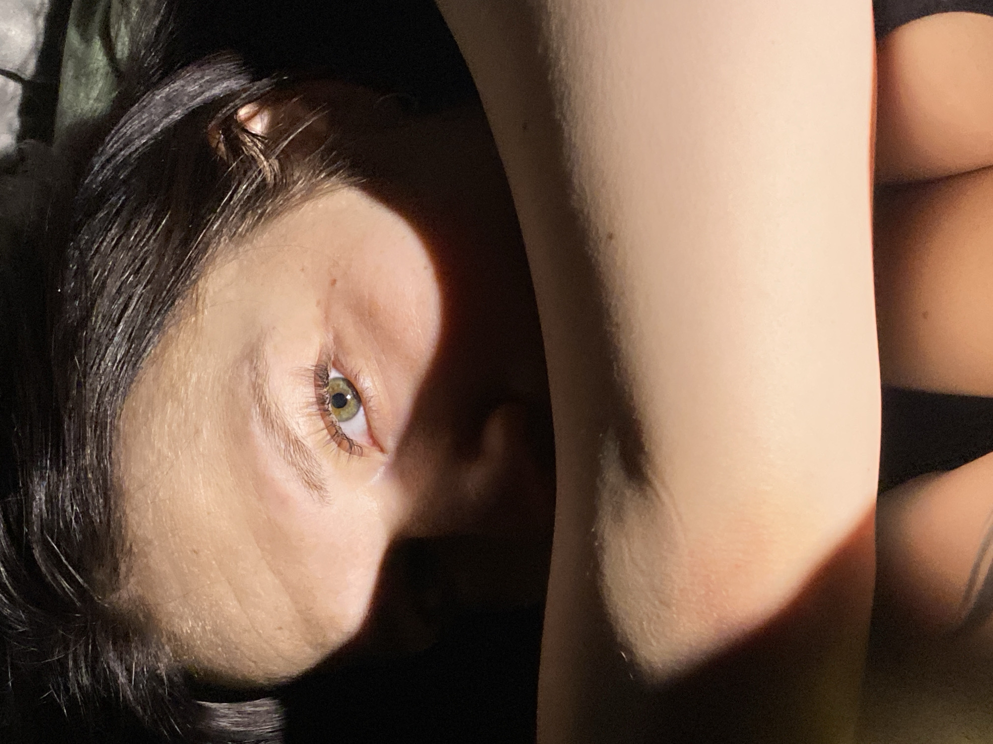 The image depicts a person with dark hair and light skin, lying down. Their face is turned towards the camera, with a green eye visible. Part of their arm or leg is also visible in the frame, and what appears to be a dark garment. The lighting is uneven, creating strong shadows across the subject. There is no information available in the image that suggests the location is Chaikovsky, Russia, or any specific location.