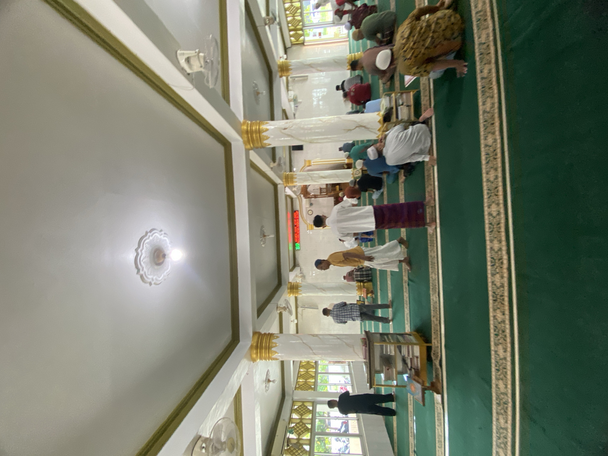 The image displays the interior of a mosque or prayer hall in Lhokseumawe, Indonesia, showing a congregational prayer in progress. The perspective is slightly elevated, looking across the length of the hall, and the image is rotated 90 degrees counter-clockwise.
Numerous men are engaged in prayer on a green carpeted floor adorned with patterned gold borders. They are observed in various postures of Salat, including prostration (sujood), standing, and kneeling. The men wear a variety of traditional Muslim attire, such as long shirts, robes, sarongs (often patterned), and prayer caps (kufis).
The architecture of the hall is notable for its several tall, decorative pillars that run down the center, featuring white marble-like finishes and ornate gold bases and capitals. The ceiling is white with gold trim, punctuated by decorative light fixtures and functional white ceiling fans. Natural light streams in through large windows equipped with gold, geometrically patterned grilles.
On a side wall, a digital prayer time display with red digits is visible. Though somewhat blurry, it clearly indicates prayer timings, with "DHUHUR 12:34" and "ASHAR 15:45" being discernible, suggesting it is daytime. A small wooden bookshelf or stand with religious texts is also present near the worshippers. The scene conveys a solemn and communal atmosphere of worship.