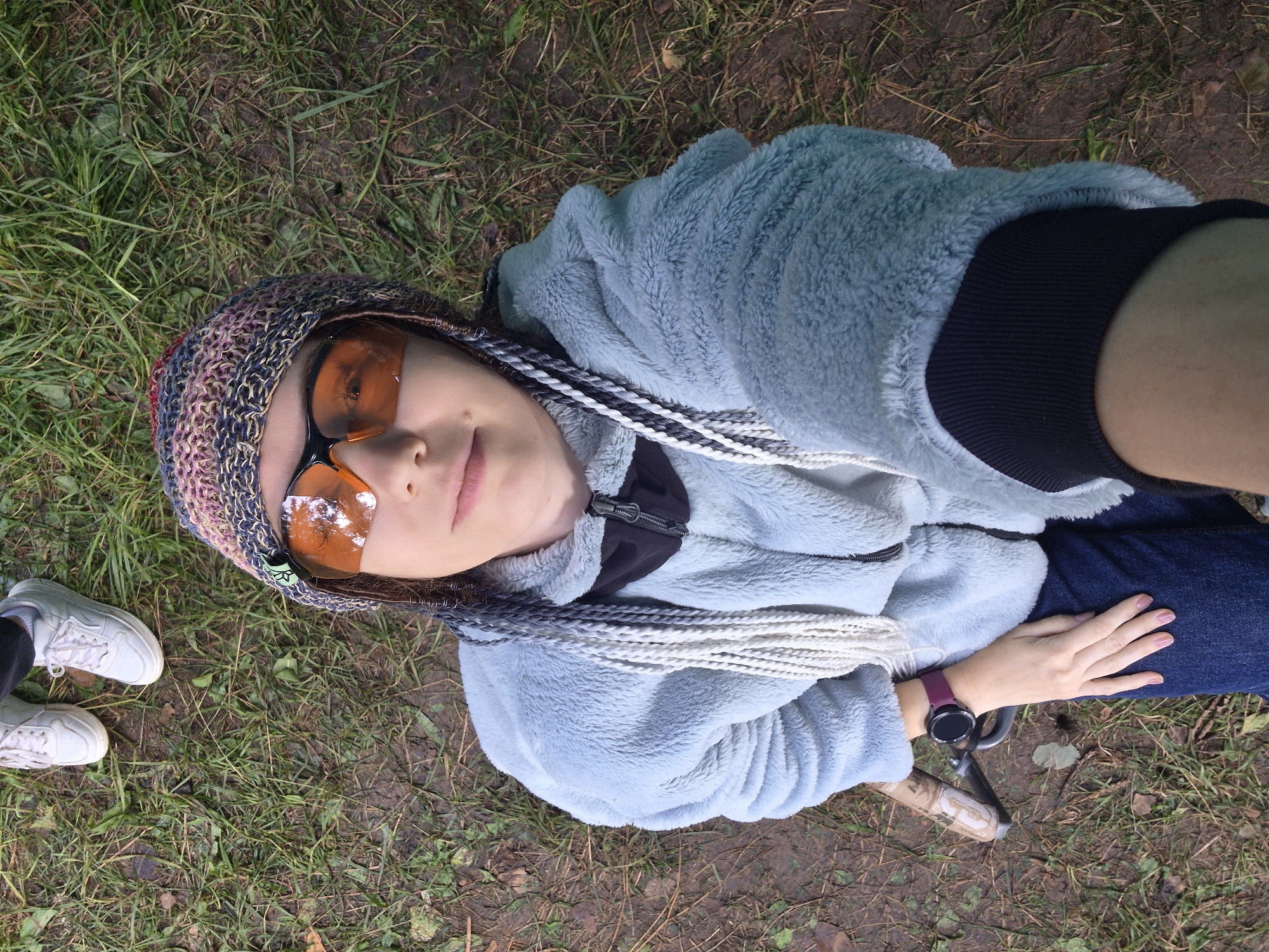 A person is taking a selfie outdoors, from a high angle, on a dirt path with scattered green grass and fallen leaves. The person is wearing a light blue fuzzy jacket, dark blue jeans, a multi-colored knitted hat, and orange-tinted glasses. They have braided hair that is partly visible. A purple smartwatch is visible on their left wrist. In the background, another person's feet wearing white sneakers are visible. The overall impression is of a casual outdoor setting, possibly a park or a wooded area. The location is not specified in the image.