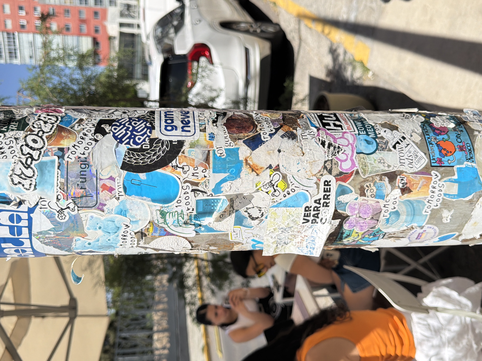 The central subject is a vertical metal pole, completely covered in a dense, overlapping collage of stickers. These stickers exhibit a wide variety of sizes, colors, and designs, ranging from vibrant graphics and logos to intricate illustrations and text.
Above the pole, the background reveals an urban environment under clear daylight. A multi-story red building with many windows stands prominently, partially obscured by green trees and a blue sky. To the right, a white car is parked on a street or sidewalk, its red taillights visible, next to a yellow-painted curb.
Below the pole, several individuals are visible in the blurred foreground, seated at tables under a large, light-colored awning or umbrella. One person wears an orange top, seen from the back, while others with dark hair are partially obscured. They appear to be engaged in an outdoor social or dining activity.
The bright sunlight and pronounced shadows indicate it is likely mid-morning or afternoon. The scene conveys a lively, everyday urban atmosphere.
Numerous texts are visible on the stickers, with OCR identifying recurrent phrases such as "TOMORROW GOODS," "Fungi GOOD," "gam neve," and "OUR COFFEE." Other notable inscriptions include "VER PARA CORRER," "Underground City Mexico," "BURGERS MEOW," "RUNA," and social media handles like "@almash" and "@Bone de Cord." Numerical sequences such as "10," "34001," and "540041" are also present. The combination of Spanish text and the "Mexico" reference on a sticker, along with the given location context of Puebla, confirms an urban Mexican setting.