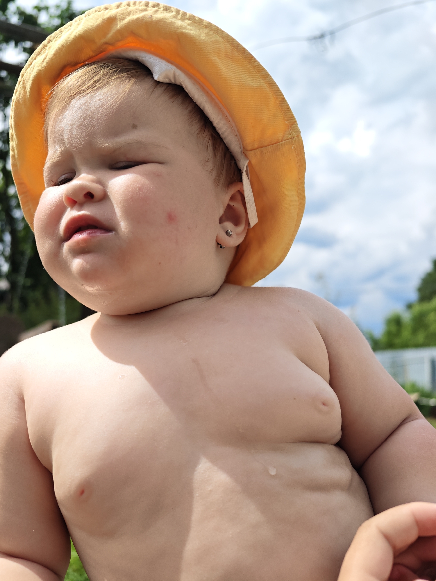 This is a close-up, low-angle outdoor photograph of a baby wearing a yellow sun hat. The baby's face is partially obscured by the hat, and they have their eyes closed, with a somewhat pensive or contented expression. A small mole is visible on their forehead, and a red mark, possibly a mosquito bite, is on their cheek. Their ears are visible, and one has a small stud earring. The baby's chest and shoulder are also visible, showing smooth, plump skin. A small red mark, perhaps another bite, is on the baby's chest. There are droplets of water on the baby's skin, suggesting they may have been in water or are sweating.
The background consists of a cloudy sky and some greenery, indicative of an outdoor setting, likely a backyard or park. The lighting suggests it is daytime, possibly late morning or afternoon, with bright sunlight filtering through the clouds. The presence of the sun hat and the baby's attire (or lack thereof, as they appear shirtless) suggest warm weather. The overall mood of the image is relaxed and candid, capturing a moment of childhood. The location context of Noviy, Russia, places the scene within that geographical area. There is no visible text in the image.
