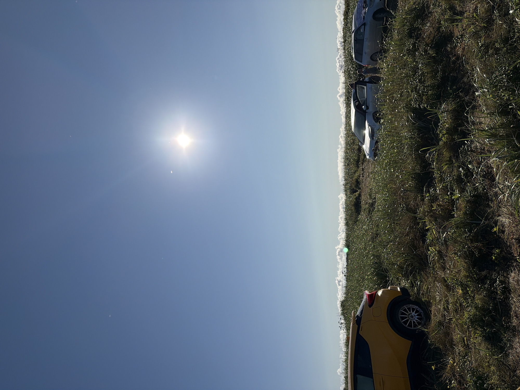 The image depicts a bright, sunny day in a rural or coastal setting, characterized by a clear blue sky with the sun prominently featured. The foreground shows a grassy field with several cars parked. A yellow SUV is on the left, and further to the right, there are at least two other cars, one appearing to be silver and another white, with people standing near them, some with car doors open. The landscape is covered in green vegetation, with hints of flowering plants or dried stalks. In the distance, a hazy line of white suggests clouds or mist, possibly over water or a lower elevation. The lighting suggests it's daytime, with the sun high in the sky. The image does not provide any specific visual cues to pinpoint the location as Ozerskoe, Russia.