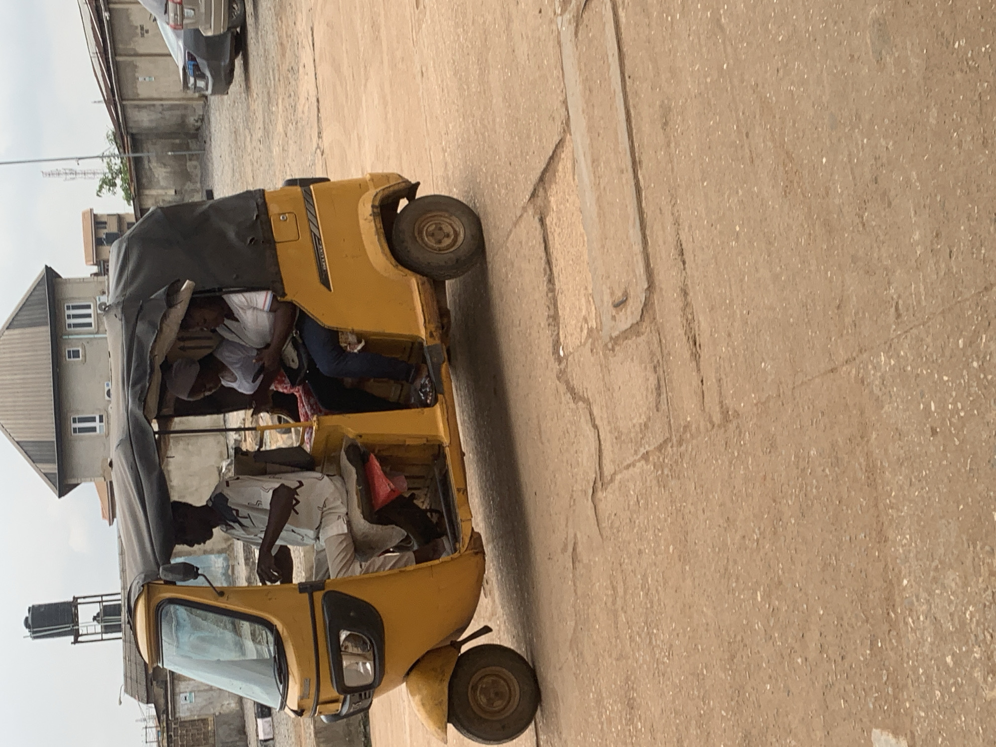 The image shows a yellow tricycle taxi, commonly known as an "okada" or "keke napep," with three occupants. A man, likely the driver, is seated in the front, operating the vehicle. Two women are seated behind him. The tricycle is traveling on a dirt road in Shagamu, Nigeria, with buildings and other vehicles visible in the background. The sky is overcast.