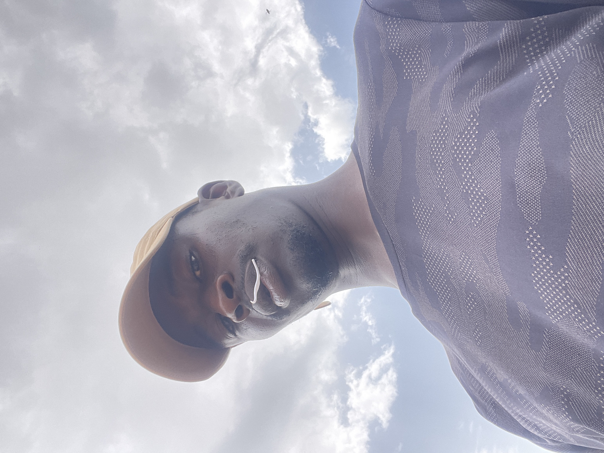 A low-angle shot shows a dark-skinned man wearing a tan baseball cap and a blue patterned shirt. He is looking down at the camera. In his left ear, there is a white wireless earbud. The background is a cloudy sky. No information from the image suggests the location is Durimi, Nigeria.