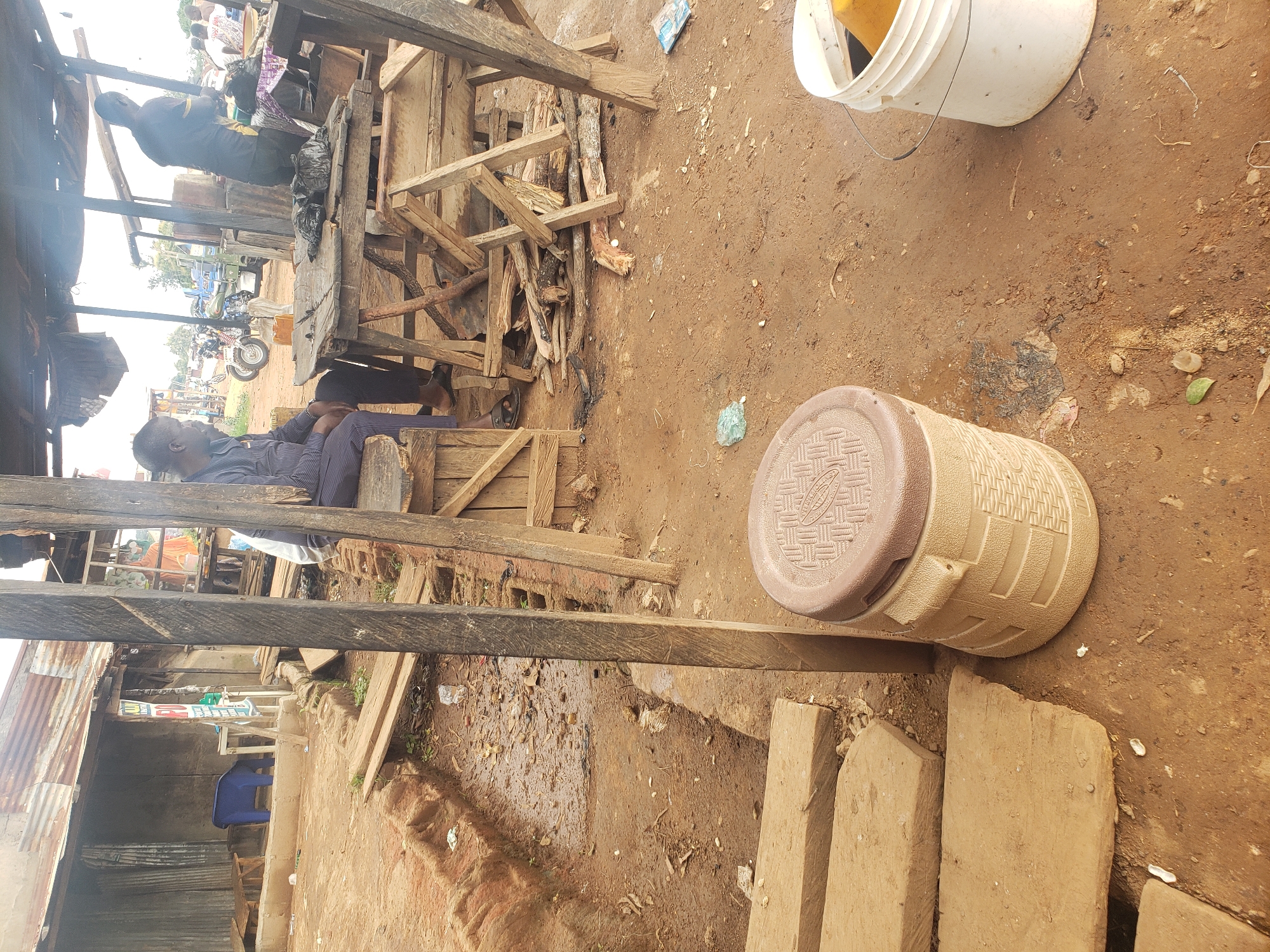This is a medium shot of an outdoor market scene in Kachia, Nigeria. The ground is dirt and appears muddy in places, with scattered debris and dry leaves. To the left, a partially constructed wooden structure with a corrugated tin roof is visible. Inside, a blue plastic chair is overturned on the ground.
In the foreground, a large, light brown cooler with a darker brown lid sits on the dirt. Several rough wooden planks are laid out like steps or a path in front of it. Further back, a wooden table supported by a-frame legs is set up under a thatched roof. Piled on the table are black plastic bags and some firewood.
Two men are present. One man, wearing a dark pinstripe suit and sandals, is seated on a wooden bench to the left, facing away from the camera. Another man, wearing a dark t-shirt and light-colored pants, stands to the right of the table, facing away from the camera and appearing to be engaged in an activity. In the background, more structures and a few motorbikes are visible, along with other indistinct figures. A white bucket containing a yellow object is to the right of the cooler. The overall impression is of a functional, perhaps temporary, marketplace.