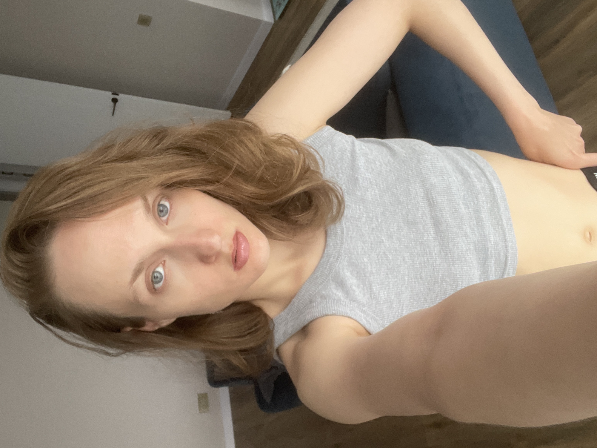 A light-skinned woman with light brown hair and blue eyes is visible. She is wearing a gray tank top and lying on a blue surface. Her location in Подшивалово, Russia, cannot be determined from the image. The background shows a wall, electrical outlets, and flooring.