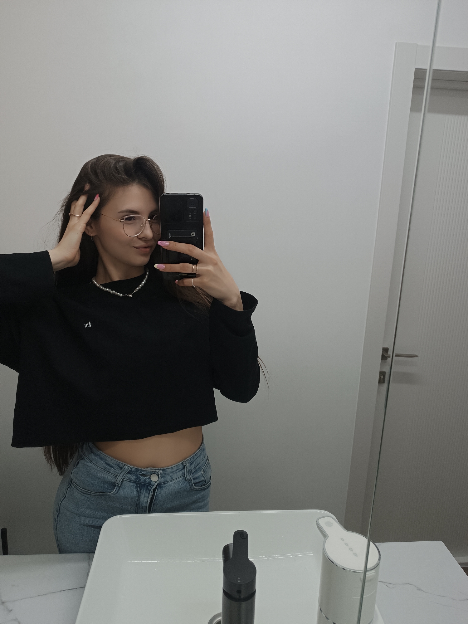 A woman is taking a selfie in a bathroom. She is wearing a black crop top, blue jeans, and glasses. She has long, brown hair and is wearing a pearl necklace. The bathroom has a white sink and a mirror. There is a door to the right of the mirror. The city and location are unknown.