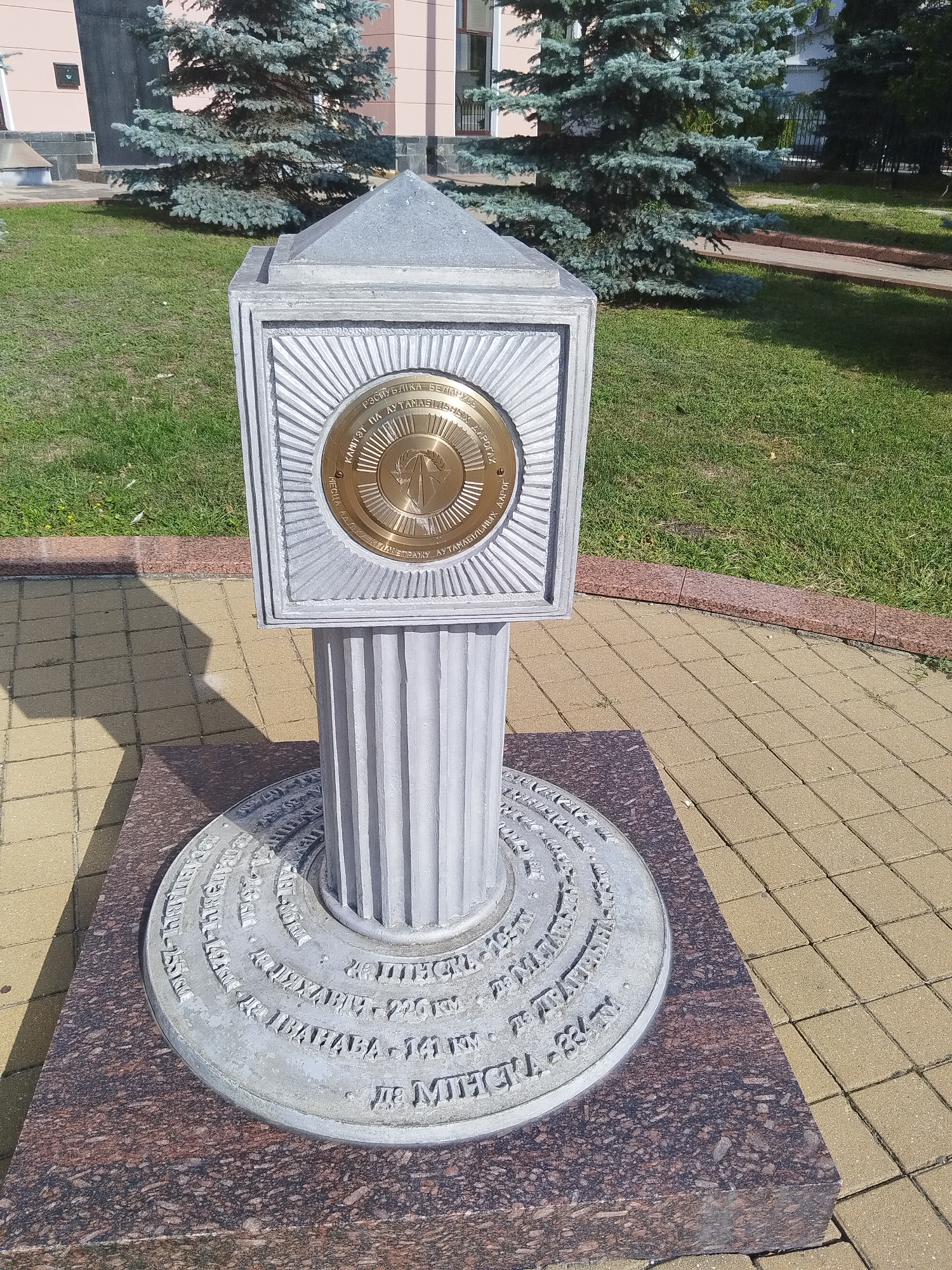 The image shows a monument located in Brest, Belarus. The monument consists of a circular base inscribed with text, a fluted column, and a square top with a circular medallion. The medallion, made of gold-colored metal, features an emblem and text in Cyrillic. The monument is situated on a paving stone area, with a lawn and trees in the background. A shadow of the monument is cast on the paving stones. The overall setting appears to be an outdoor public space.