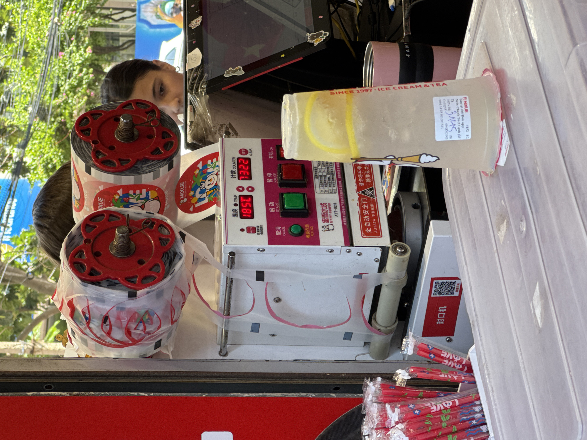 This image, captured in Nha Trang Ward, Vietnam, depicts a beverage service counter. In the foreground, a clear plastic cup filled with a pale yellow liquid, ice, and several lemon slices rests on a light grey, textured surface. The cup features vertical text "SINCE 1997 ICE CREAM & TEA" and a sticker identifying it as "MIXUE" brand "Nước Chanh Tươi Lạnh (M)" (Lemon Fresh Water, Medium).
To the left of the cup, a container holds multiple individually wrapped red plastic straws, many printed with "LOVE" and leaf patterns. Dominating the left-center of the midground is a white and grey automatic cup-sealing machine, branded "MIXUE" and "ATT-B2H". Two red spools on top hold transparent plastic film rolls adorned with red "MIXUE" branding and cartoon characters, with the film threaded into the machine. The control panel displays a "温度 TEMP" (Temperature) of "185C" and a "计数 COUNTER" (Count) of "1322". It features green "启动 START" and "卷膜点动 ROLLING" buttons, and red "暂停 PAUSE", "开机 ON", and "取消 CANCEL" buttons. A red label below displays "封口机" (Sealing Machine) and a QR code.
To the right of the cup, a black computer monitor, decorated with stickers including a possible flag, is angled towards the right. A pink and black insulated tumbler is partially visible behind the cup. In the background, a young adult female with dark hair is partially visible behind the equipment, looking downwards. Beyond the counter area, through an opening, green foliage, trees, blue and white signs, and a partially obscured vehicle are visible, suggesting an outdoor environment.