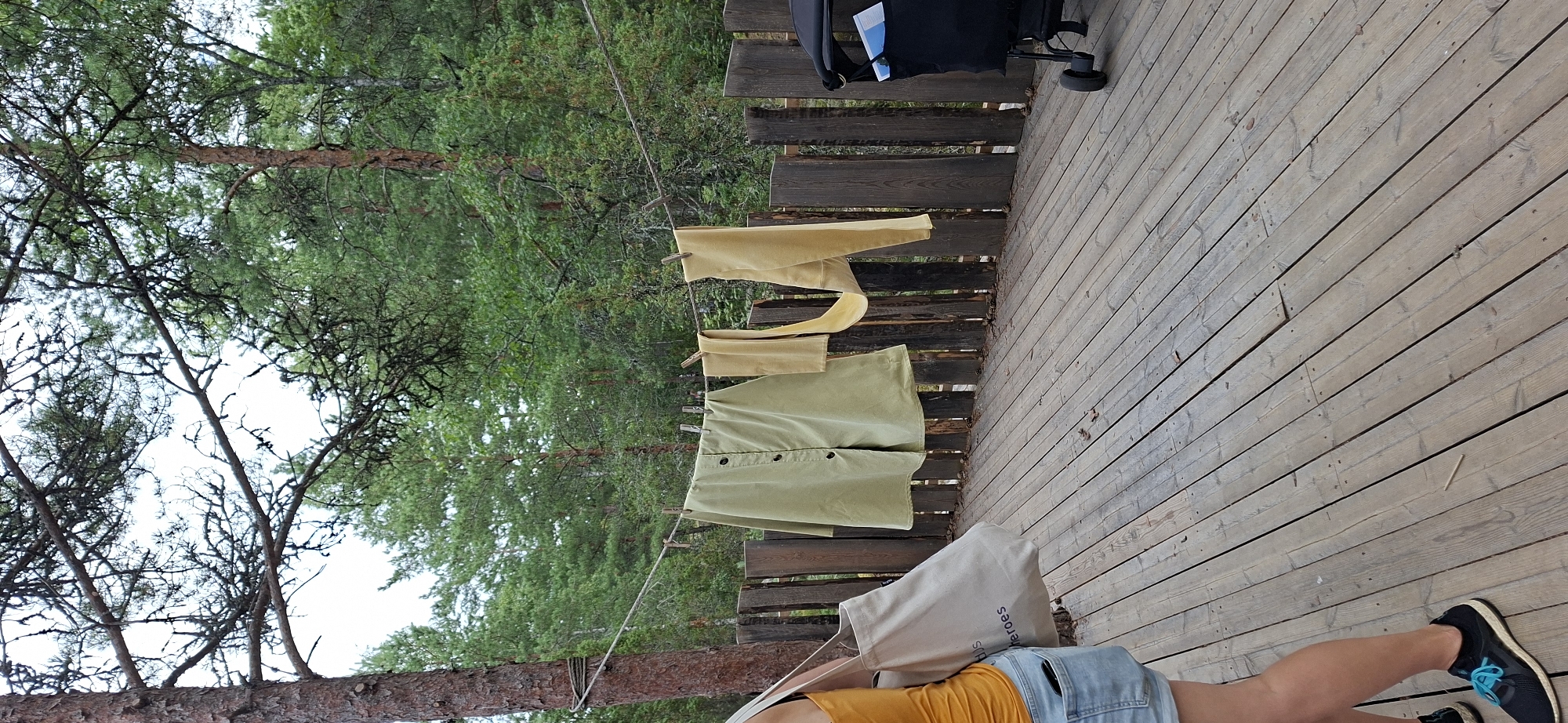 In Naantali, Finland, a wooden deck area is shown. A person, partially visible, stands on the left, wearing a yellow tank top and denim shorts, carrying a canvas tote bag. Clothes are hanging on a clothesline stretched between trees and a rustic wooden fence. The clothes include a light green button-up garment and two yellow pieces of fabric, possibly pants or a long skirt. A black stroller with a blue booklet is parked on the right side of the deck. The background consists of lush green trees, suggesting a natural or park-like setting. The overall scene captures a moment of outdoor laundry drying amidst a tranquil, wooded environment.