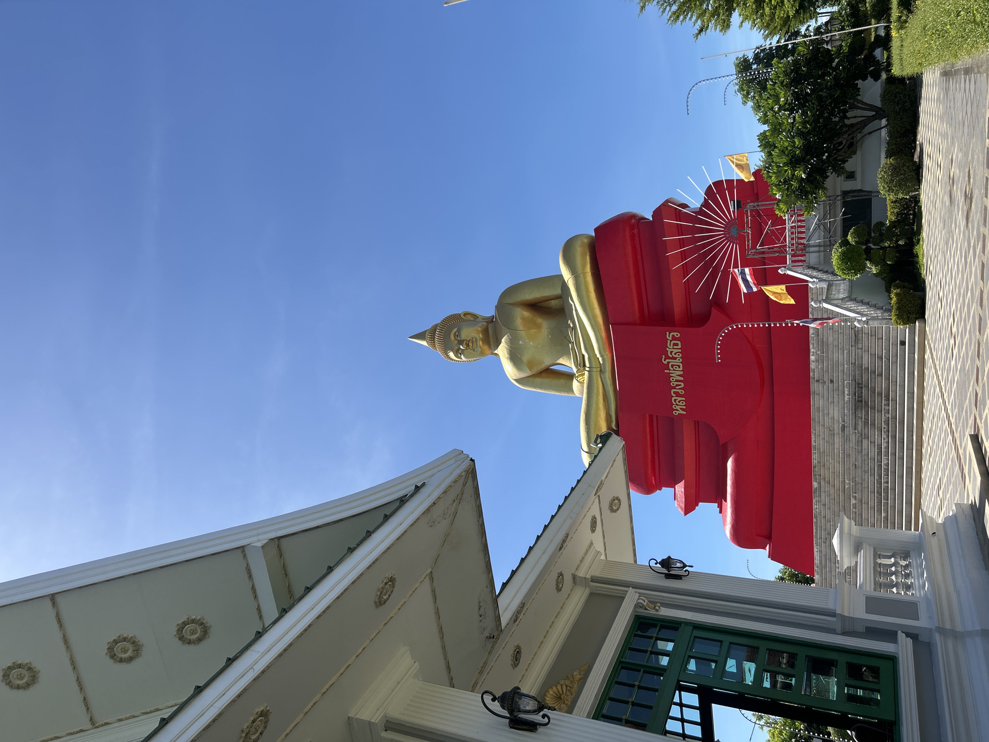 An outdoor, eye-level, slightly low-angle, full shot captures a bright, sunny day in Thai Ko Subdistrict, Thailand. The scene features a large, gold-colored Buddha statue seated in a meditative pose atop a vibrant red, multi-tiered pedestal. The Buddha statue is prominent in the upper right quadrant of the image, with its gold features sharply defined against a clear, bright blue sky. The red pedestal is adorned with Thai script in gold lettering and is accessible by a white-railed staircase. Two yellow flags and one Thai flag are visible on the stairs, suggesting a place of cultural or religious significance.
In the lower left quadrant, the corner of a white building with an ornate, overhanging roof is visible. The building features a green-framed door with multiple panes and traditional outdoor lighting fixtures attached to its white pillars. The building's roof is decorated with circular floral patterns. The ground in front of the building and leading towards the stairs is paved with a mosaic-like pattern of light and dark gray square tiles. Lush green trees and manicured shrubs are present to the right of the pedestal, framing the view and indicating a well-maintained outdoor space. The overall impression is one of reverence and tranquil beauty under a clear sky.