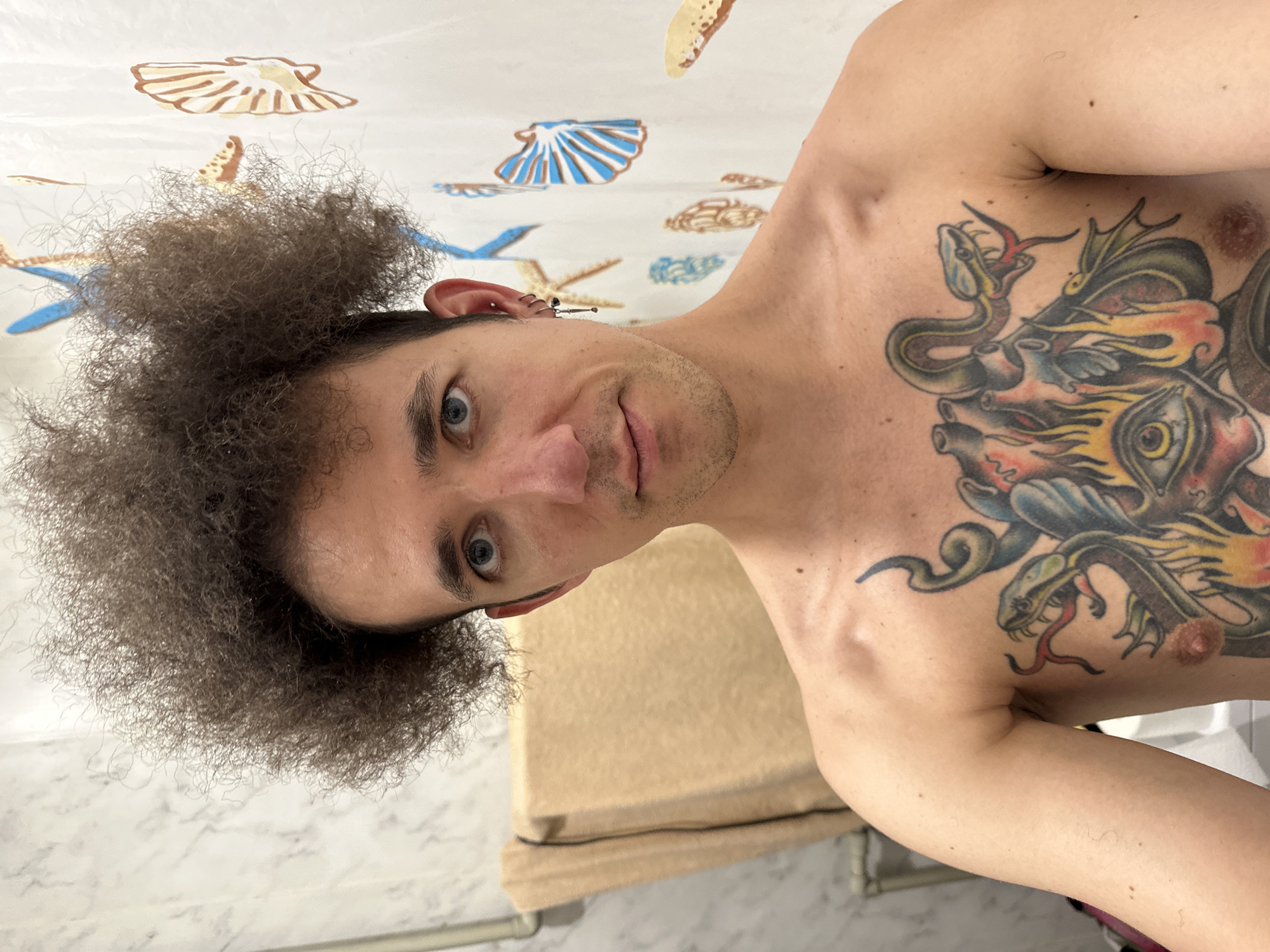A man with an afro, blue eyes, and a large chest tattoo is taking a selfie in a bathroom. He is shirtless. The background shows a marble-tiled wall, a towel rack with a tan towel, and a shower curtain with a seashell pattern. The man is wearing earrings. The image was taken in Saratov, Russia.