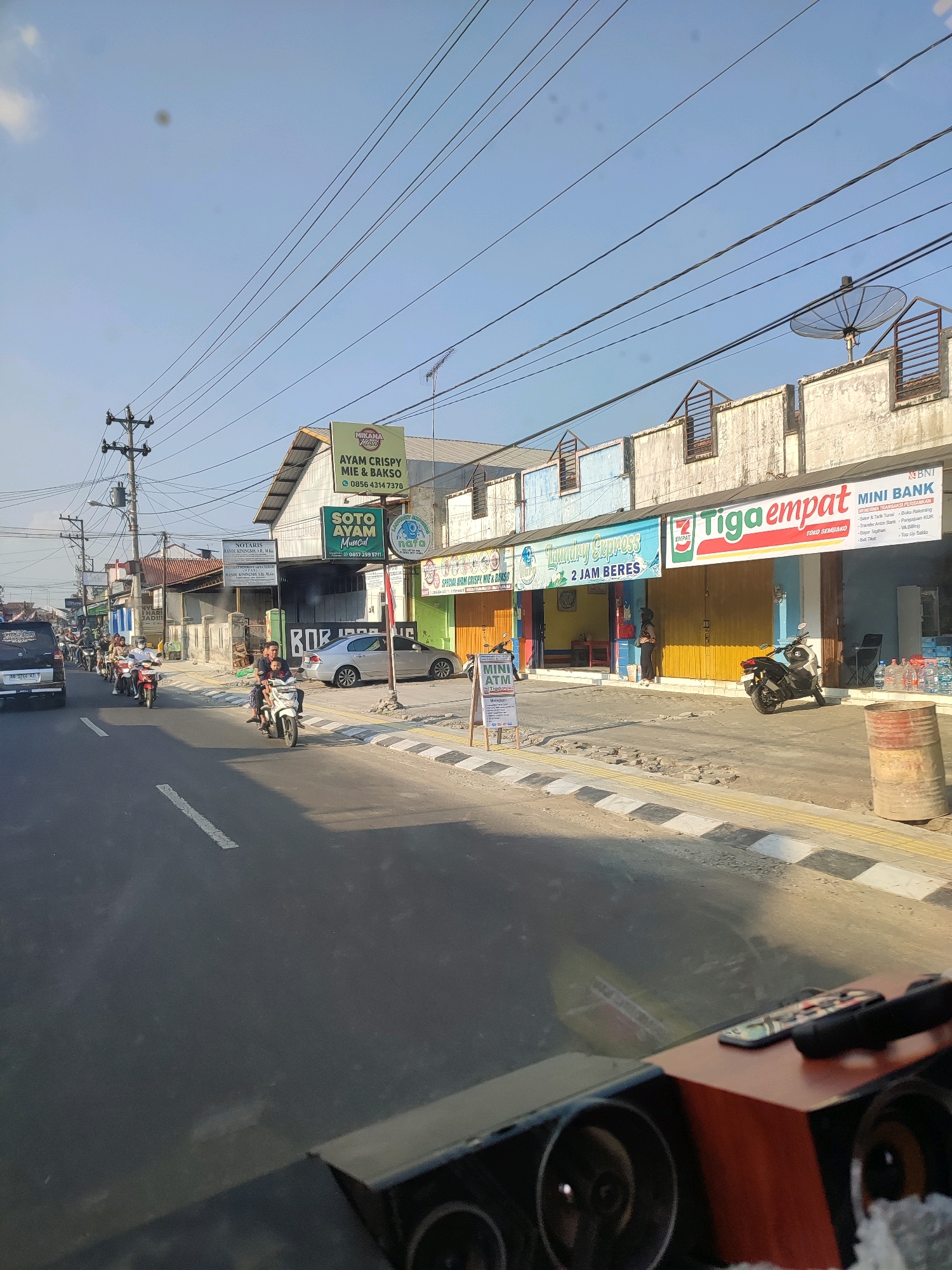 The image depicts a street scene in Magelang, Indonesia, under a clear sky. A road with white lane markings stretches into the distance, bordered by a sidewalk. Several motorcycles are visible on the road, with some carrying passengers, indicating ongoing traffic. Buildings line the right side of the street, housing various businesses, including a "MIKANA KRESS" eatery, "Laundry Express," a "Tiga Empat TOKO SEMBAKO," and a "BNI MINI BANK." A satellite dish is mounted on the roof of one of the buildings. People are seen interacting with these establishments, with one person standing in front of the laundry service and another near the mini-bank. Further down the street, more shops and a parked silver car are present. Electrical wires crisscross the sky above the buildings. The overall atmosphere suggests a typical day in a bustling commercial area.