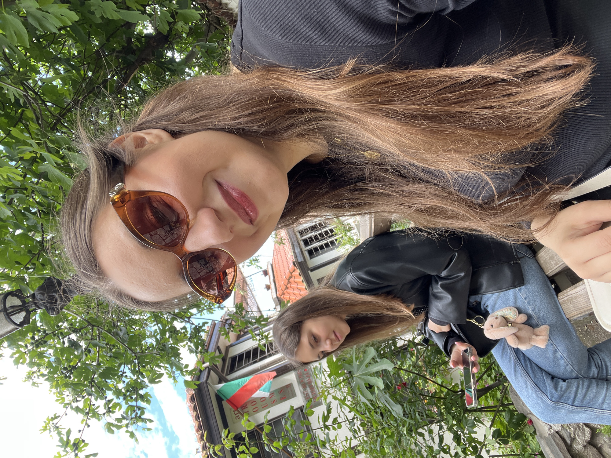 A woman is seen in a photo taken outdoors, at what appears to be a high angle. She is wearing sunglasses and has long, light brown hair. She is wearing a dark top. Another woman is seated on a bench, wearing a black leather jacket and jeans. She is holding a mobile phone. A plush bunny is on her jeans. A Bulgarian flag is visible on a building. The scene is in the city of Plovdiv, Bulgaria.