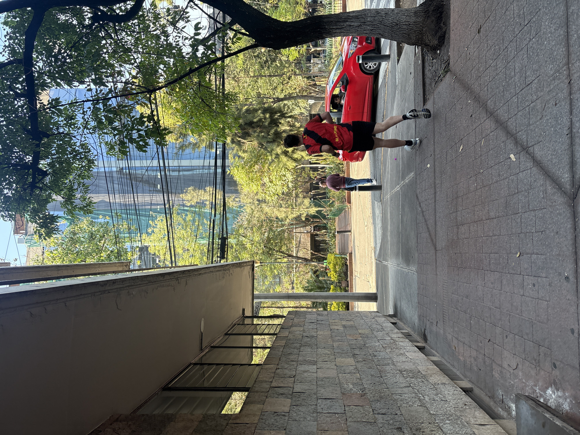A daytime scene on a sidewalk in Mexico City, Mexico. A man in a red and black sports shirt and black shorts is jogging. A red car is parked near a tree. Another person is standing on the sidewalk nearby. The architecture includes a building with a stone facade and windows. Above, trees obscure the view of a tall building. Power lines stretch across the sky.