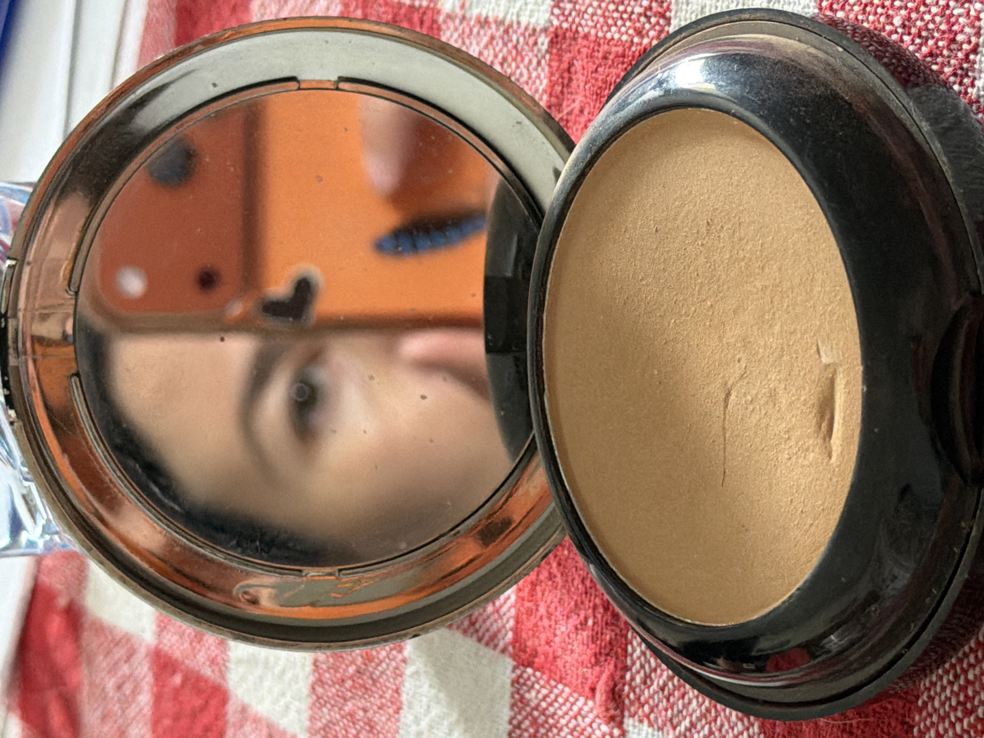 The image shows an open compact of face powder, with a mirror reflecting part of a face. The powder is a beige color and the compact has a black casing. The mirror is round and reflects an eye and part of the face. The background is a red and white checkered surface. There is no indication of the location being Tbilisi, Georgia in the image.