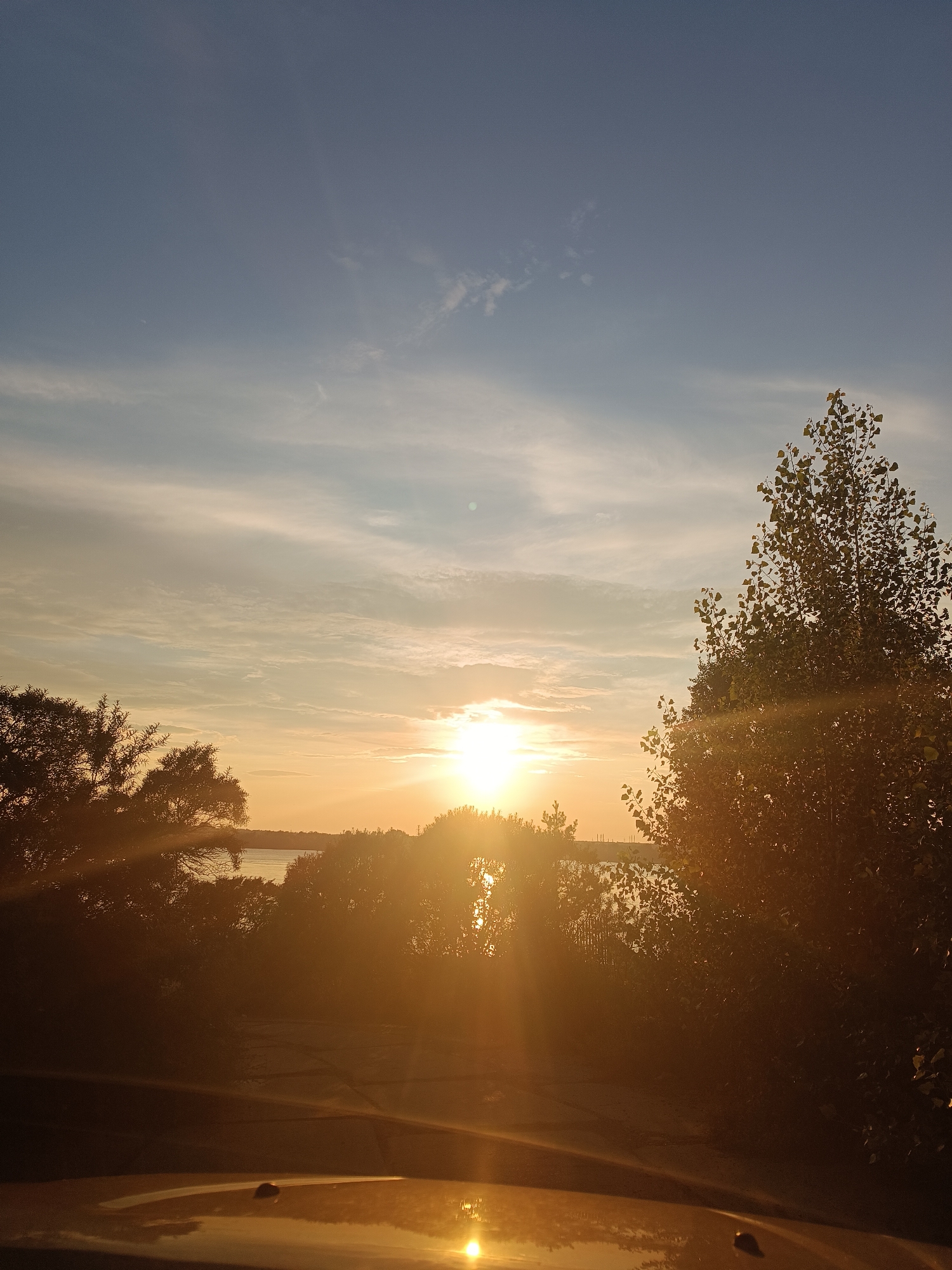 This photograph captures a serene sunset over a body of water, likely the Kama River, near Chaikovsky, Russia. The sky is a gradient of blue and orange, with wispy clouds illuminated by the setting sun. Silhouetted trees frame the scene, their leaves catching the warm light. In the foreground, the hood of a vehicle reflects the vibrant colors of the sunset. The overall impression is one of tranquility and natural beauty.