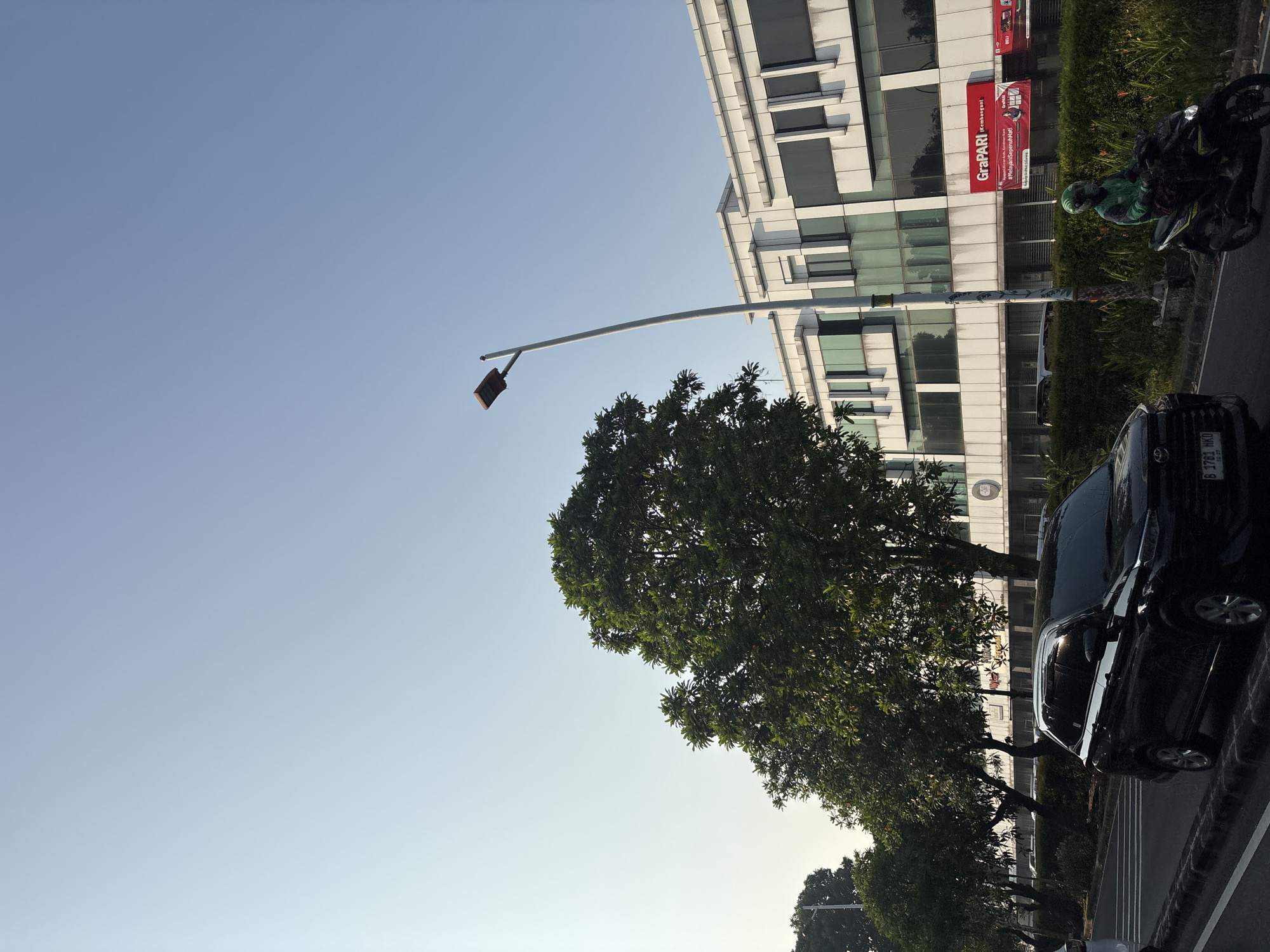 The image shows a street scene in West Jakarta, Indonesia, on a clear, sunny day. A modern, white building with large windows lines the right side of the street, featuring a red sign that reads "GraPARI" and "KEMBANGAN," indicating a location in the Kembangan area of West Jakarta. A tall streetlamp with a rectangular light fixture stands in front of the building.
On the street, a black SUV is parked on the left, and a motorcycle with a rider wearing a green helmet and jacket is moving to the right, in the same direction as the SUV. The road is paved, with white lane markings. Lush green trees with dense foliage line the street between the road and the building, casting some shadows. The sky above is a clear, pale blue.