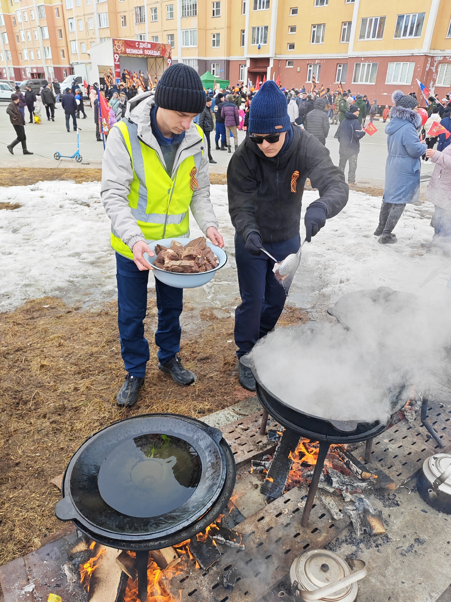 Two men are cooking food outdoors near a crowd of people. The location is Ugolnye Kopi, Russia, possibly during a celebration or public event. A man wearing a yellow reflective vest is holding a light blue bowl containing pieces of cooked meat. The other man, wearing a blue beanie and sunglasses, is using a slotted spoon to put the meat into a large cauldron filled with steaming broth. The cauldron sits over a wood fire. Another cauldron with a lid is next to it. There are tea kettles on the ground nearby. People stand in the background near a stage decorated with celebratory flags, and flags are being waved by those in the crowd. The ground is partly covered with melting snow and patches of dry grass. There is a multi-story building in the background.