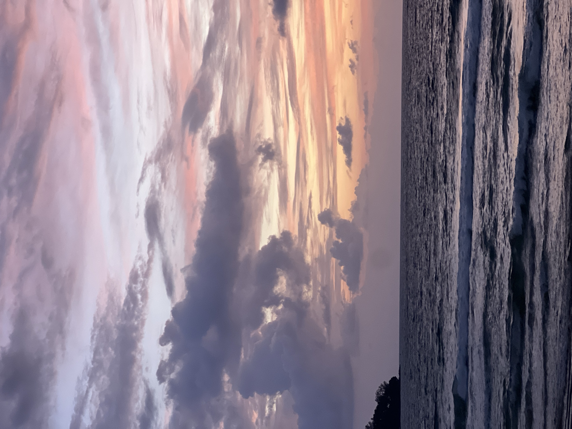 A wide shot captures a serene seascape at dusk, likely at Surin Beach in Choeng Thale, Thailand. The sky is a canvas of soft hues, transitioning from deep purples and grays of scattered clouds in the upper left to delicate pinks and oranges on the right, hinting at the recently set sun. The ocean stretches horizontally across the frame, its surface rippled with gentle waves that break in frothy white lines, creating a textured foreground. The water itself is a dark, moody blue, reflecting the dimming light. On the far left horizon, a dark, tree-covered headland juts out into the sea, silhouetted against the twilight sky. No people or vessels are visible in the scene. The overall atmosphere is one of tranquility and natural beauty.